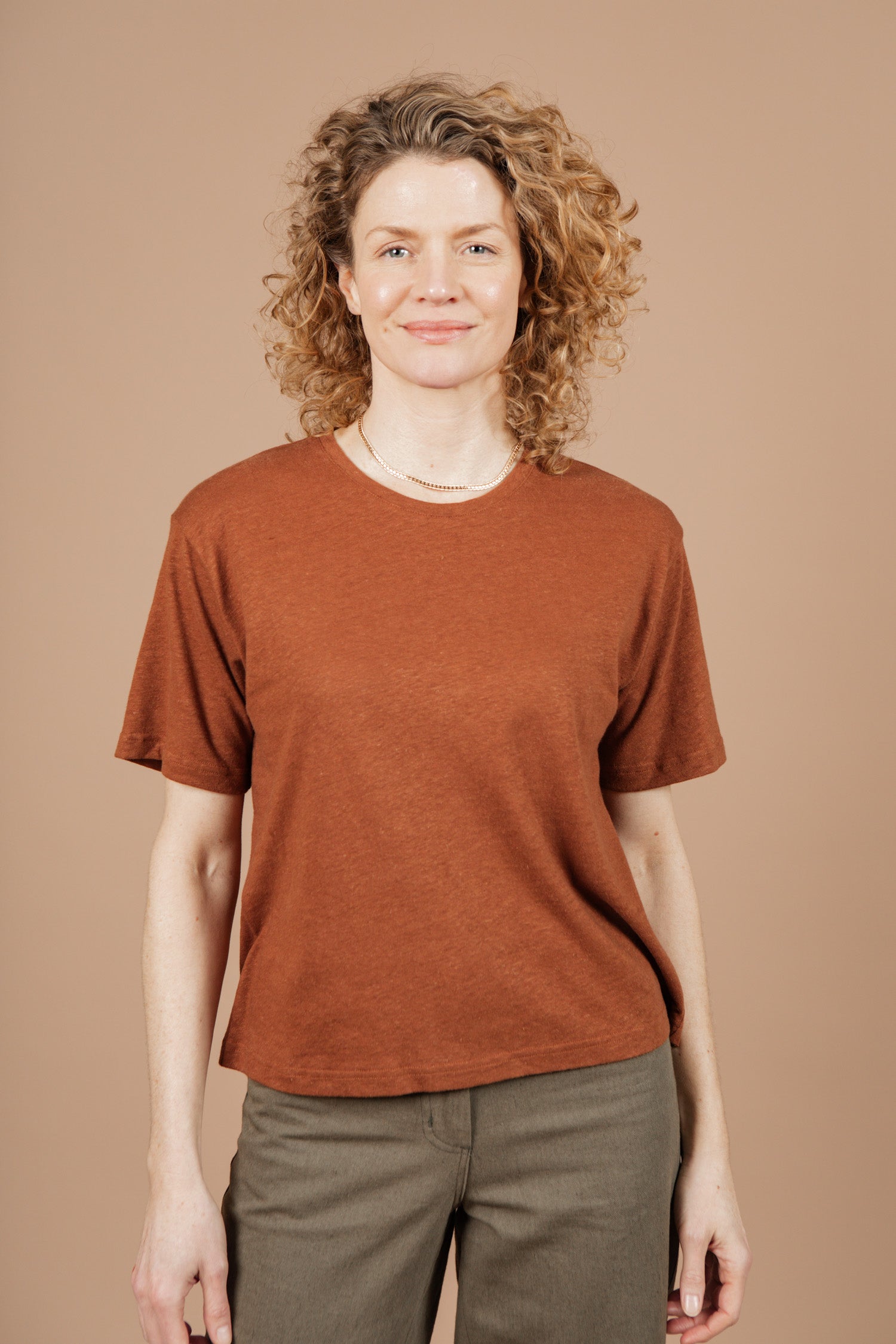 A person with curly, shoulder-length hair smiles while standing against a neutral beige background. They're wearing Bridge & Burn's Organic Hemp Boxy Tee in Burnt Umber and olive green pants.