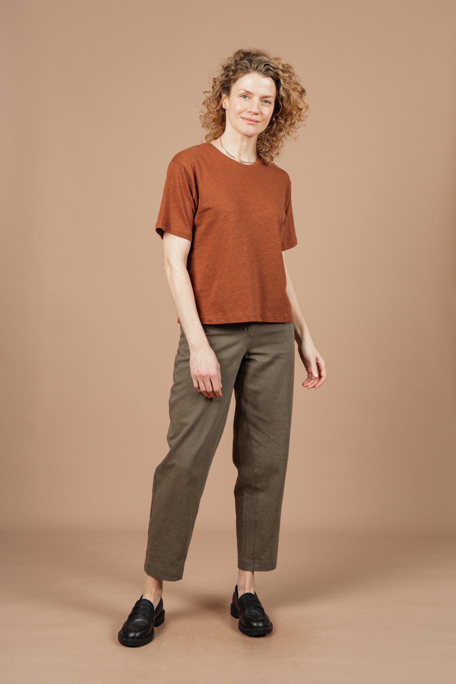 A person with curly hair stands against a beige background, wearing Bridge & Burn's Organic Hemp Boxy Tee in Burnt Umber, paired with olive green pants and black loafers, looking at the camera with a slight smile.