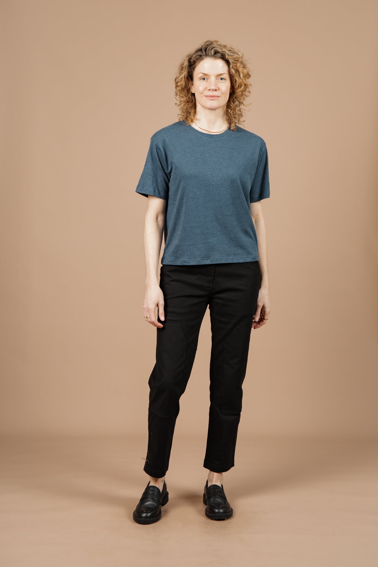 A person with curly blonde hair wears the Bridge & Burn Organic Hemp Boxy Tee in Storm Blue, paired with black pants and black shoes, standing facing forward against a plain beige background.