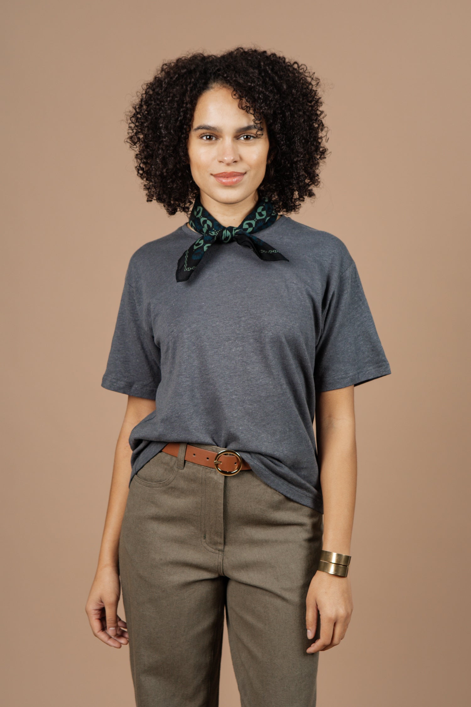 A person with curly hair wears Bridge & Burn's Organic Hemp Boxy Tee in Slate Grey, styled with olive pants, a brown belt, green patterned scarf, and gold bracelet, standing against a plain beige background.