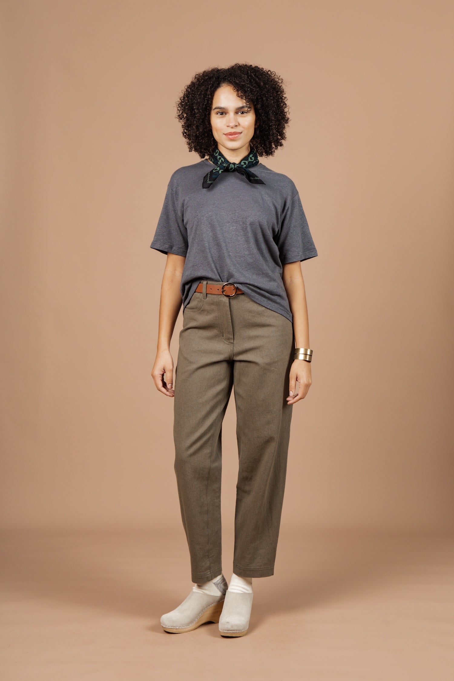 A woman with curly hair stands against a beige background, wearing the Bridge & Burn Organic Hemp Boxy Tee in Slate Grey, olive green pants with a brown belt, white shoes, a green patterned neck scarf, and a gold bracelet.