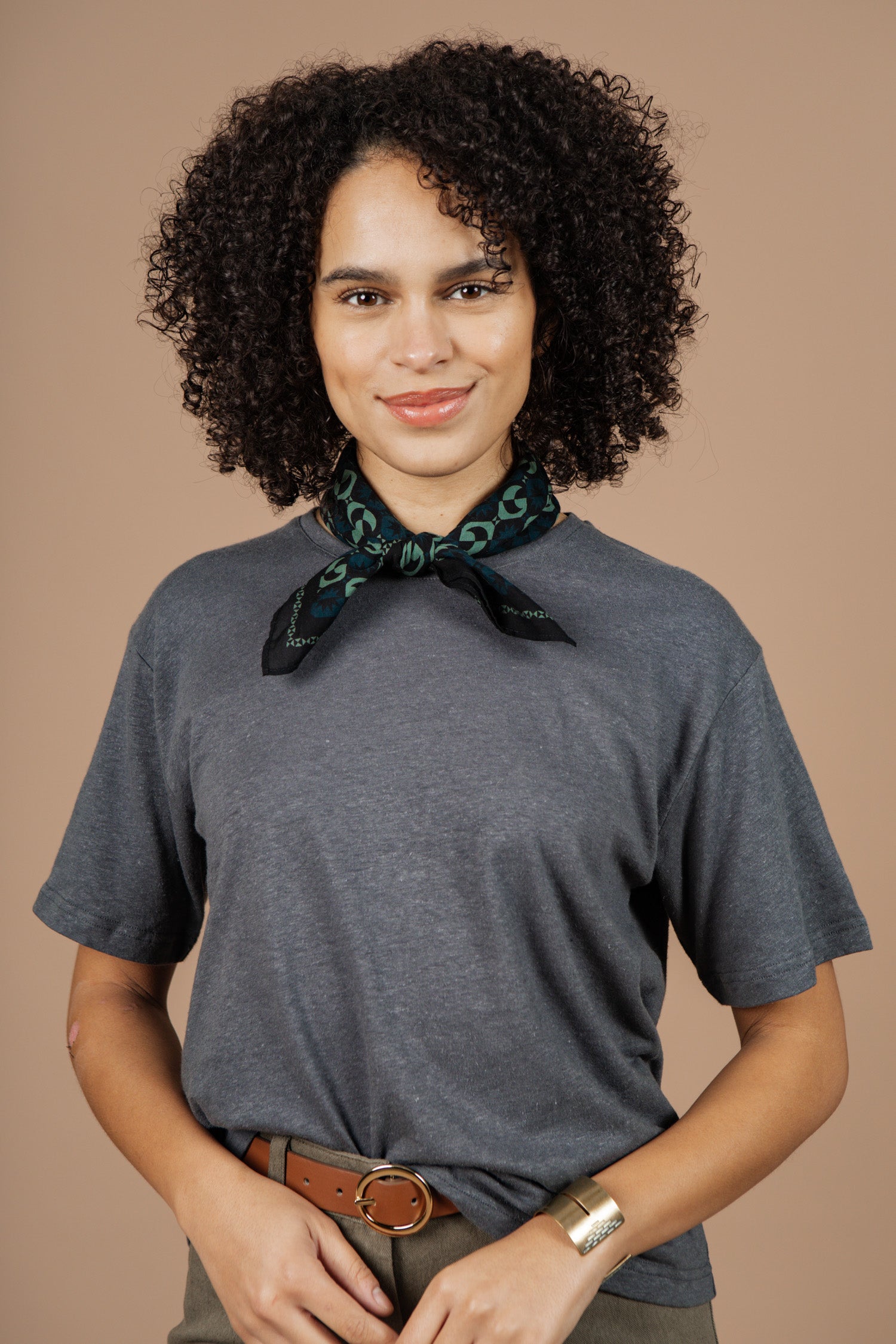 A woman with curly hair, wearing the Bridge & Burn Organic Hemp Boxy Tee in Slate Grey and a patterned scarf, brown belted pants, and a gold bracelet, stands against a beige background and smiles at the camera.