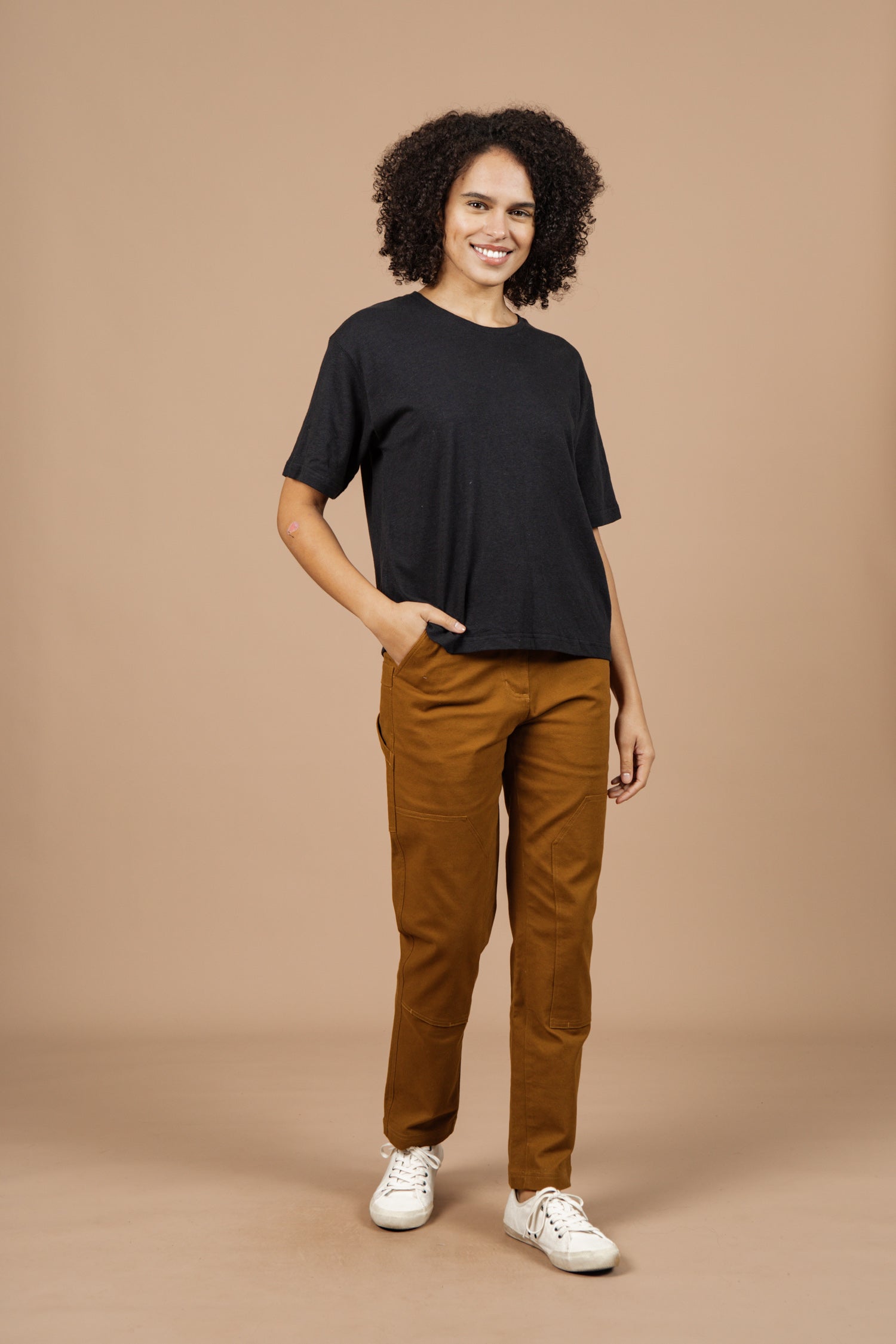 A person with curly hair is smiling, wearing the Bridge & Burn Organic Hemp Boxy Tee in Washed Black, brown pants, and white sneakers with one hand in their pocket against a beige background.