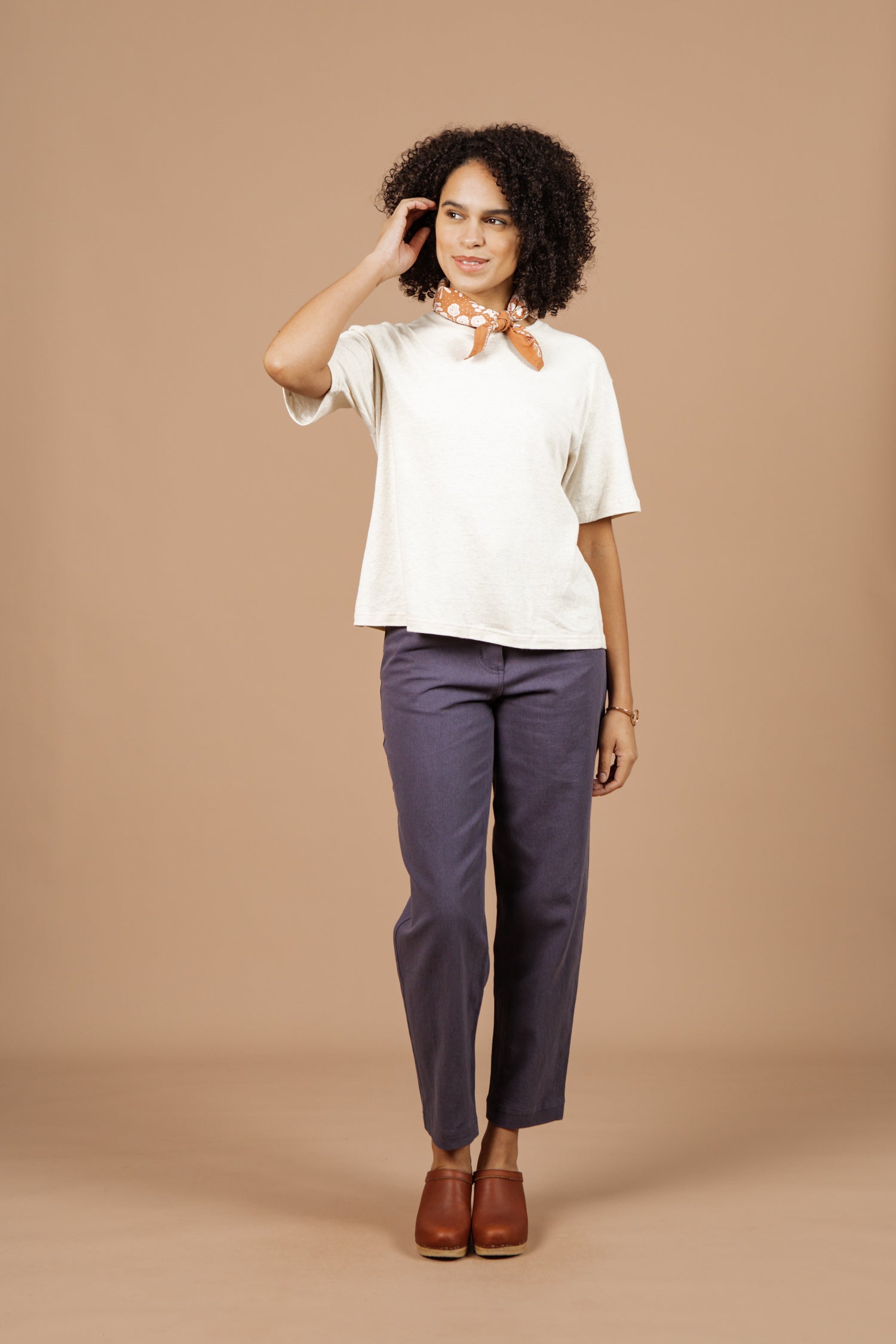 A woman with curly hair stands against a beige background, smiling as she gently touches her hair. She wears the Bridge & Burn Organic Hemp Boxy Tee in Ivory, paired with blue pants and brown accessories for an effortlessly chic look.