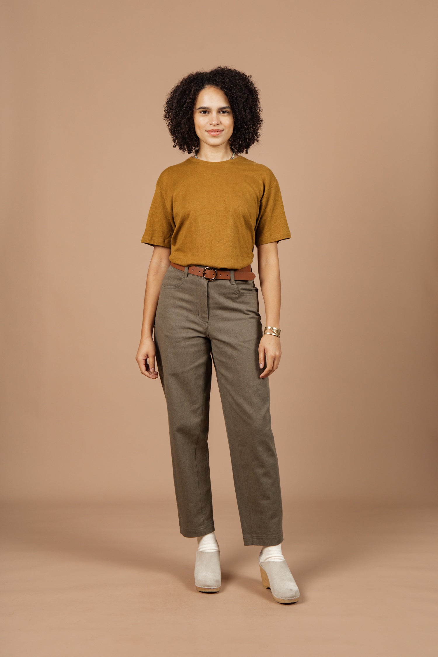 A woman with curly hair stands against a beige background, wearing the Bridge & Burn Organic Hemp Boxy Tee in ochre, high-waisted brown belted pants, white heeled shoes, and a watch. She smiles slightly and faces forward.