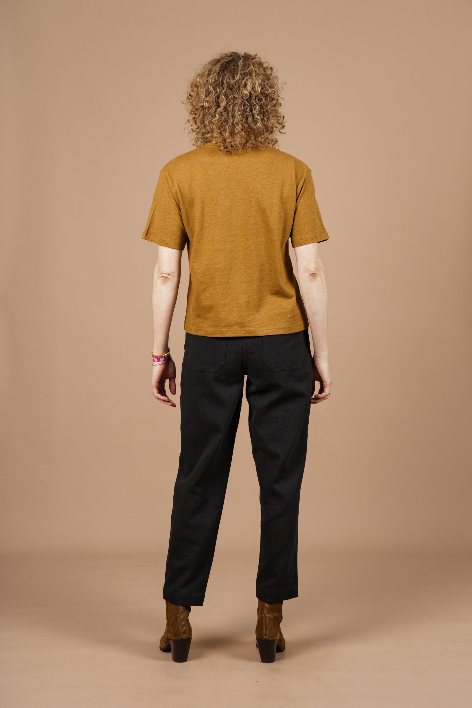 A person with curly hair faces away, wearing the Bridge & Burn Organic Hemp Boxy Tee in Ochre with black pants and brown heeled shoes, standing against a plain beige background.