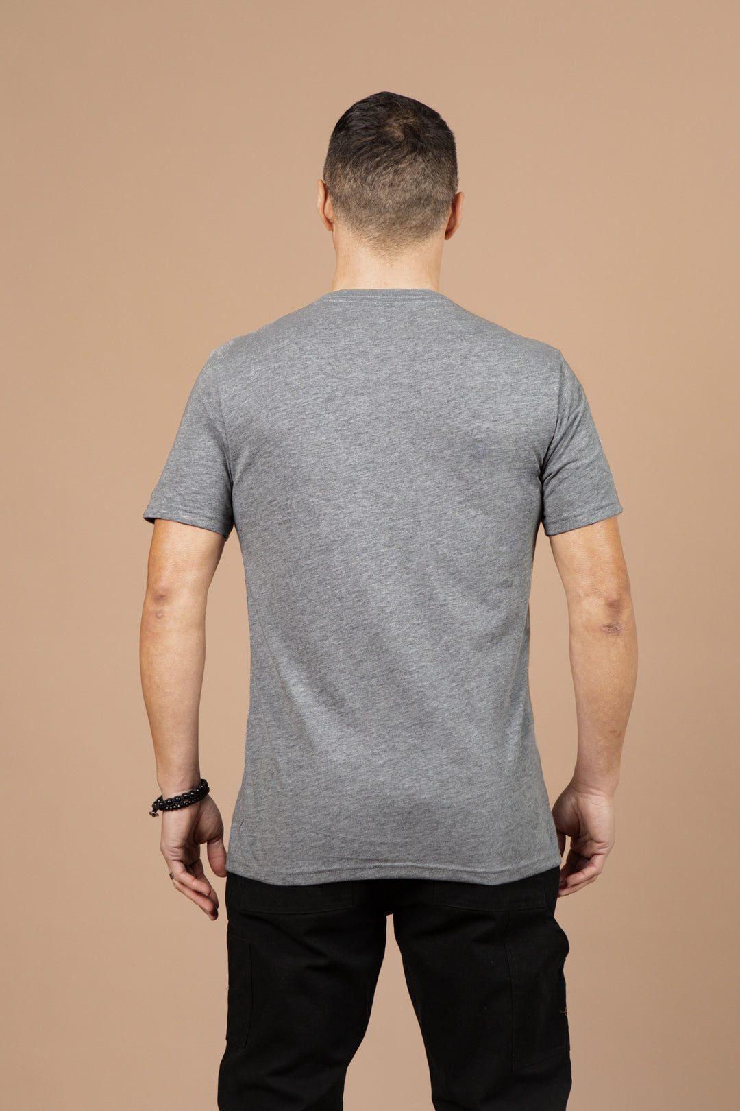 A man stands with his back to the camera, wearing Bridge & Burn’s Men's Grown Tee in grey, made from recycled polyester and organic cotton, paired with black pants against a plain beige background.