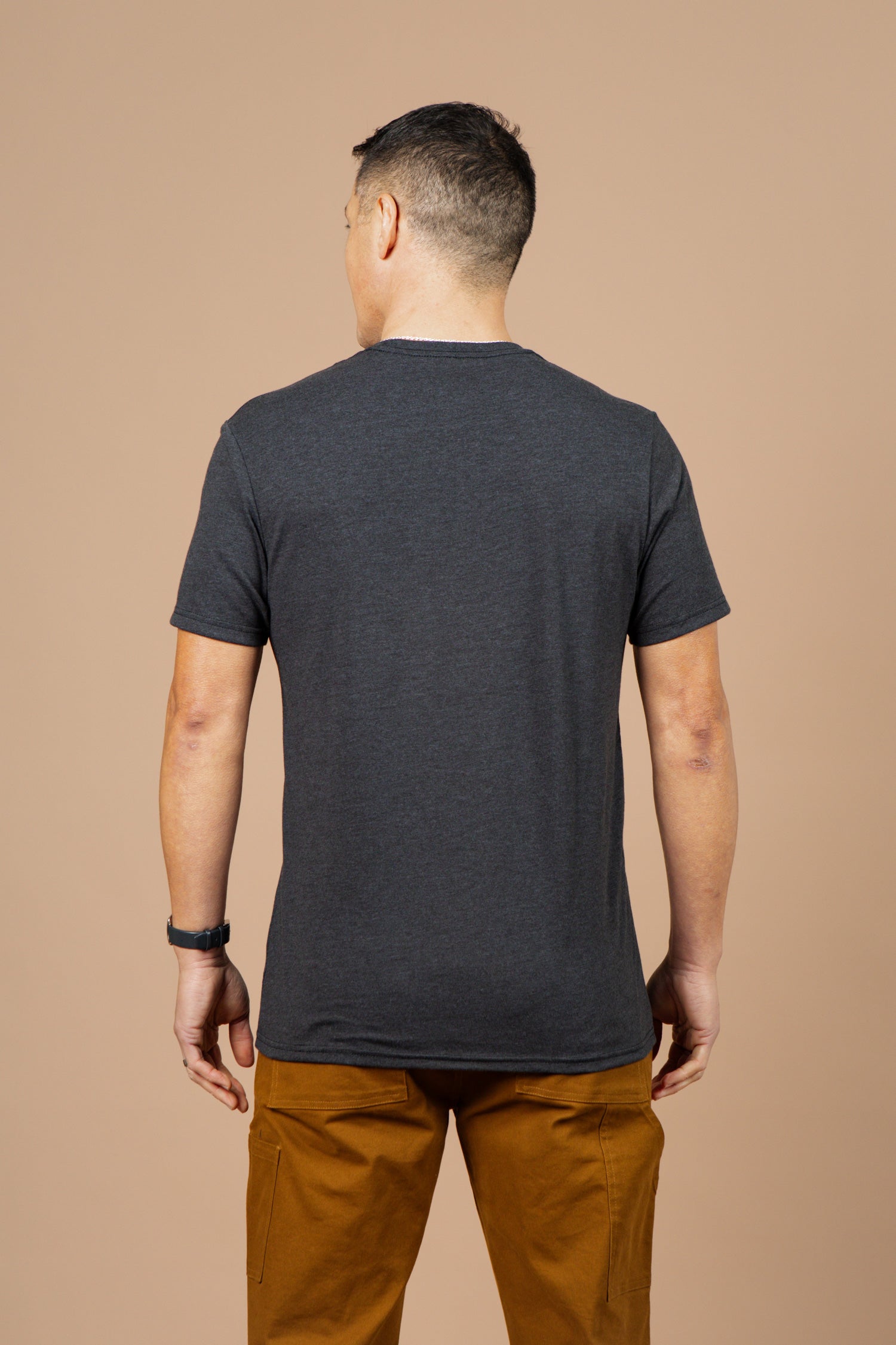A man faces away from the camera, wearing the Bridge & Burn Men's Mycology Tee in black and light brown pants, set against a plain beige background.