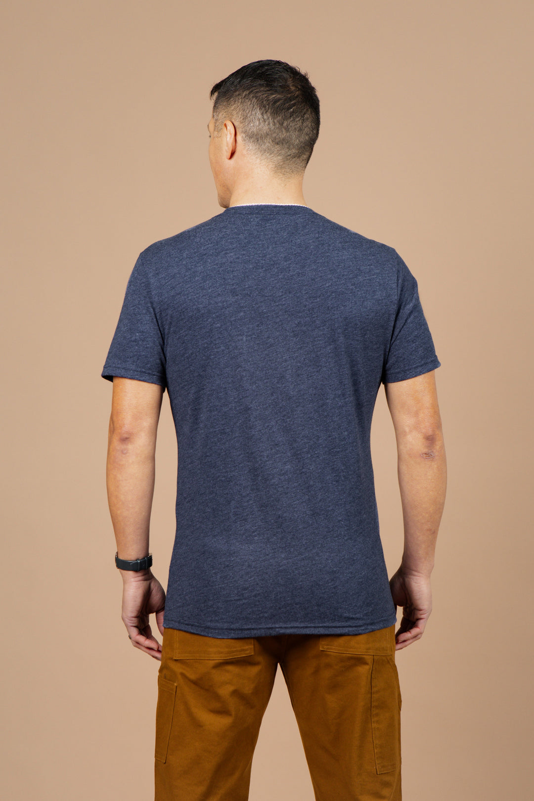 A man faces away from the camera, wearing the Bridge & Burn Men's Peace in Plants Tee in navy with light brown pants, set against a plain beige background.
