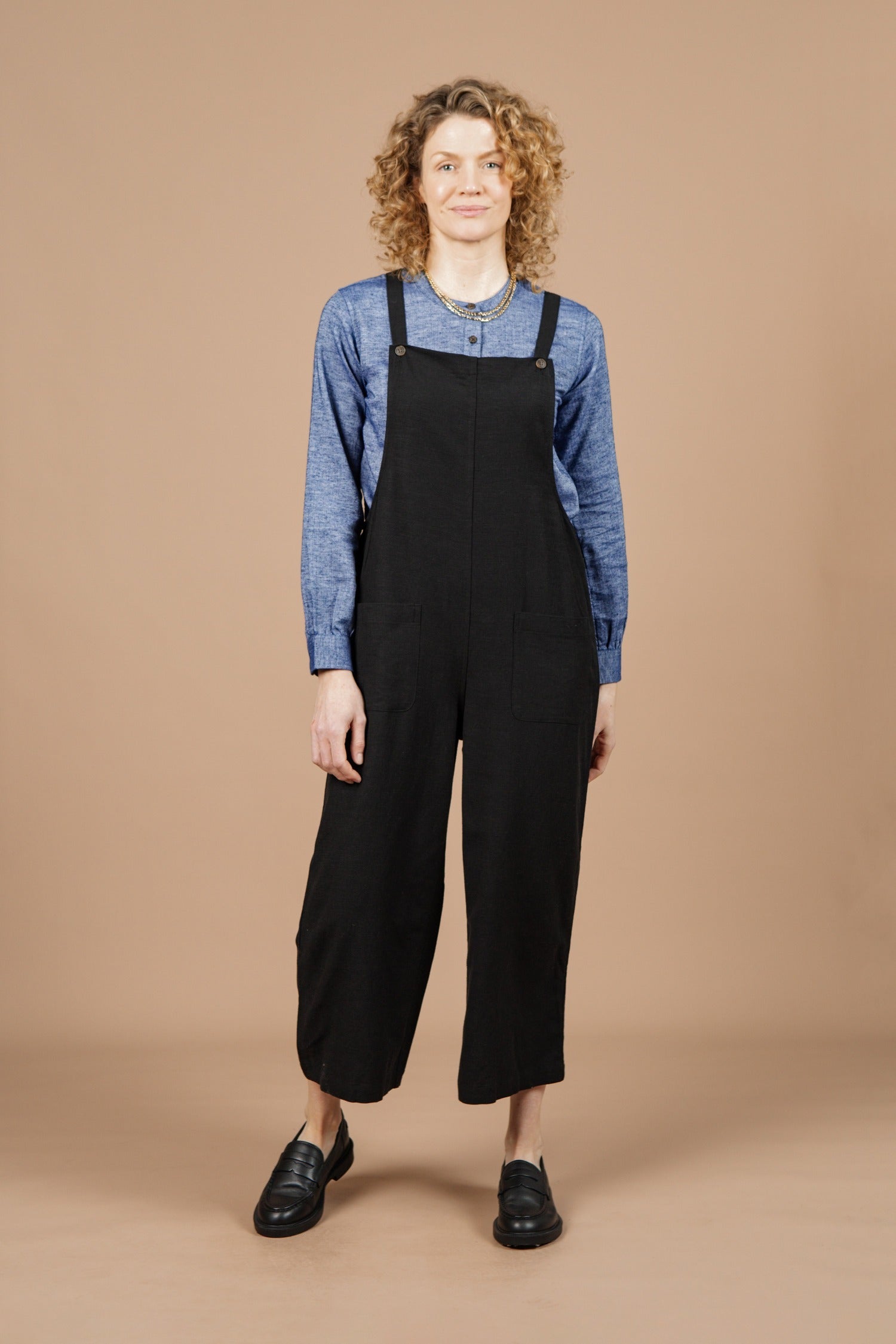 A person with curly hair smiles at the camera against a beige background, wearing Bridge & Burn's Edie Overall in Black—a wide-leg, adjustable-strap style crafted from a soft tencel-cotton blend—paired with a blue long-sleeve shirt and black shoes.