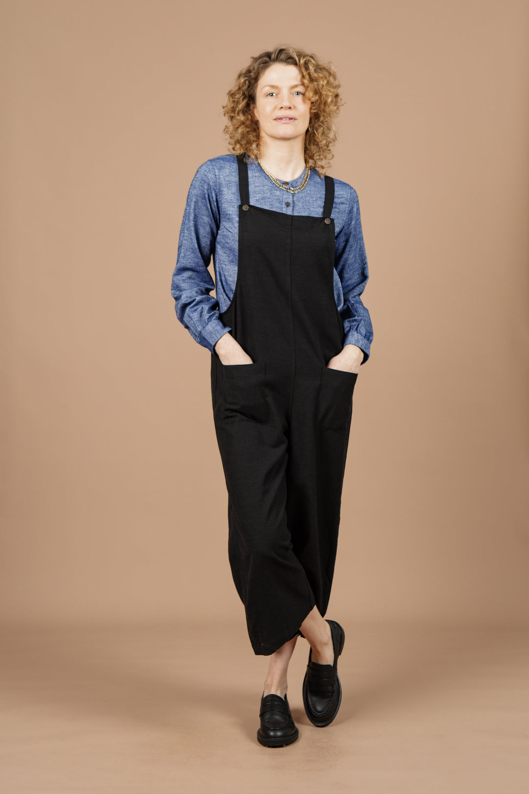 A person with curly hair wears the Bridge & Burn Edie Overall in black paired with a blue long-sleeve shirt and black shoes, standing with hands in pockets against a beige background.