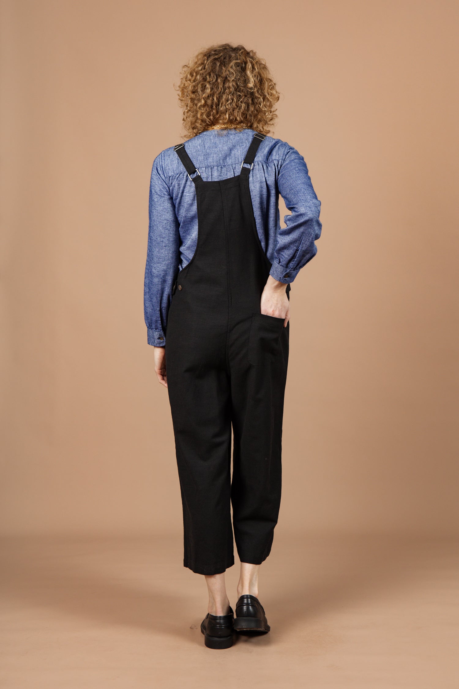 A person with curly hair, seen from behind, wears a blue long-sleeve shirt and Bridge & Burn's Edie Overall in black with adjustable straps, standing against a plain beige background.