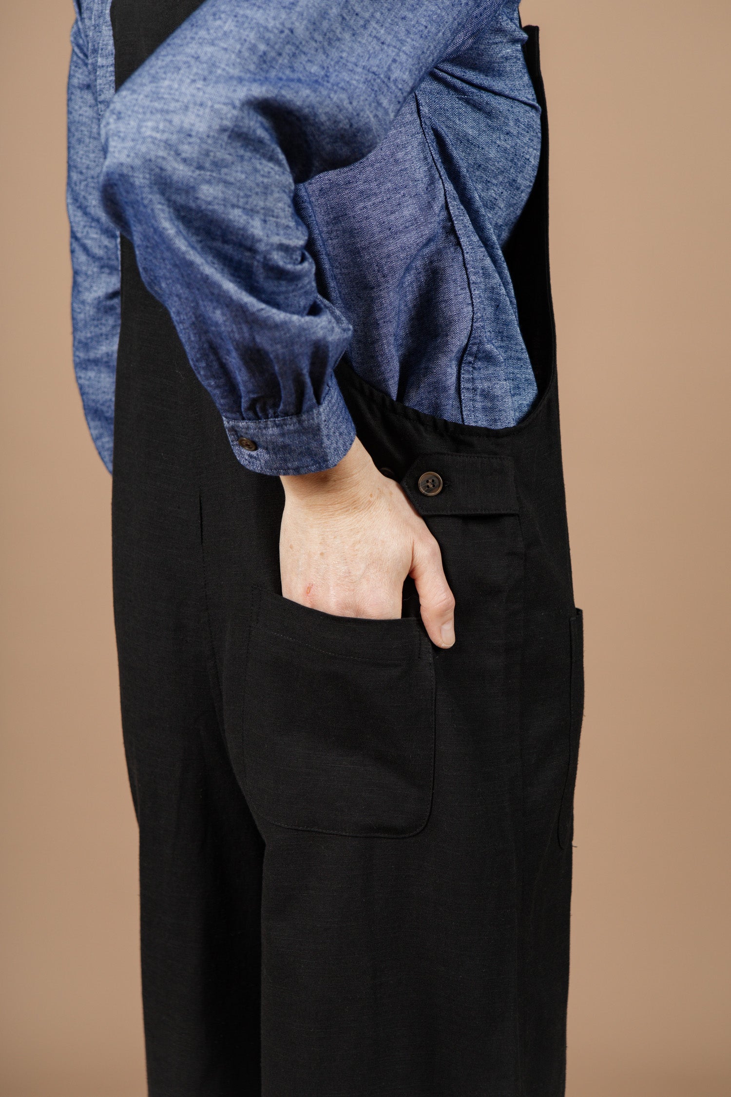 A person wearing a blue long-sleeve shirt and the Bridge & Burn Edie Overall in Black with adjustable straps stands with their hand in the pocket against a plain beige background.