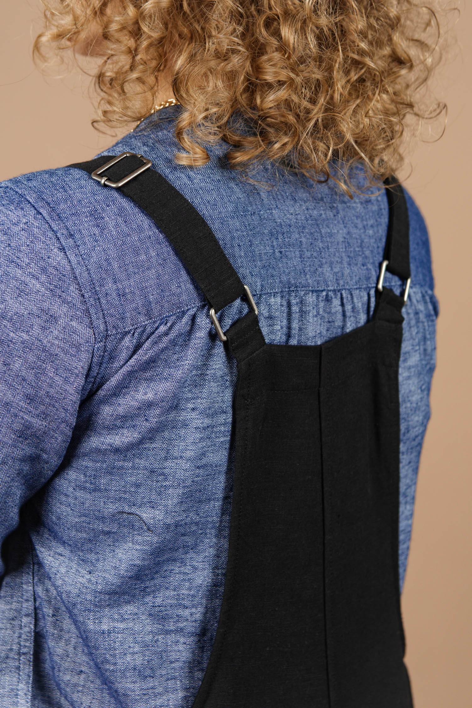 A close-up of a person with curly hair wearing the Bridge & Burn Edie Overall in black with adjustable straps and a blue denim shirt, photographed from behind against a beige background.