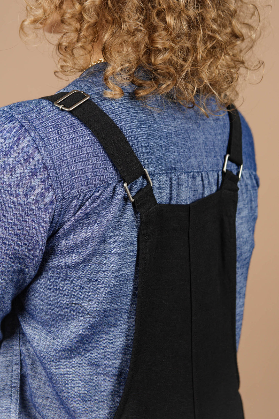 A close-up of a person with curly hair wearing the Bridge & Burn Edie Overall in black with adjustable straps and a blue denim shirt, photographed from behind against a beige background.