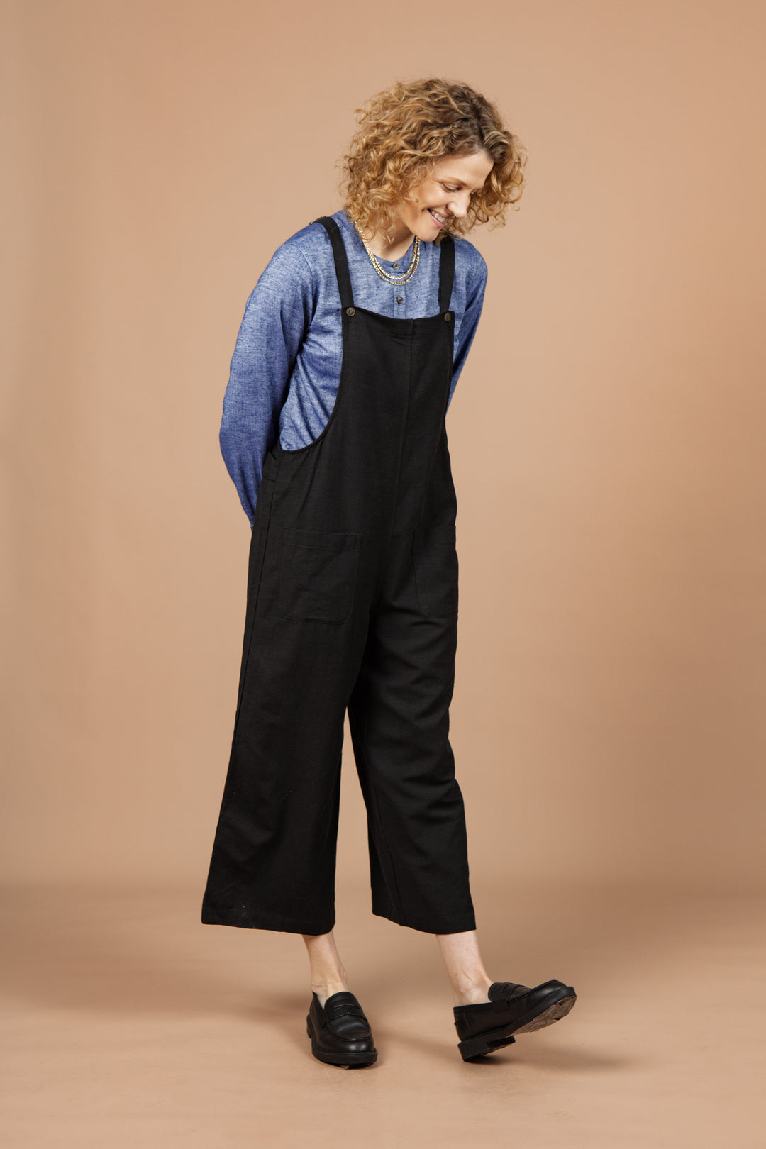 A person with curly hair wears the Bridge & Burn Edie Overall in Black over a blue long-sleeve shirt and black slip-on shoes, standing against a beige background while looking down and smiling.