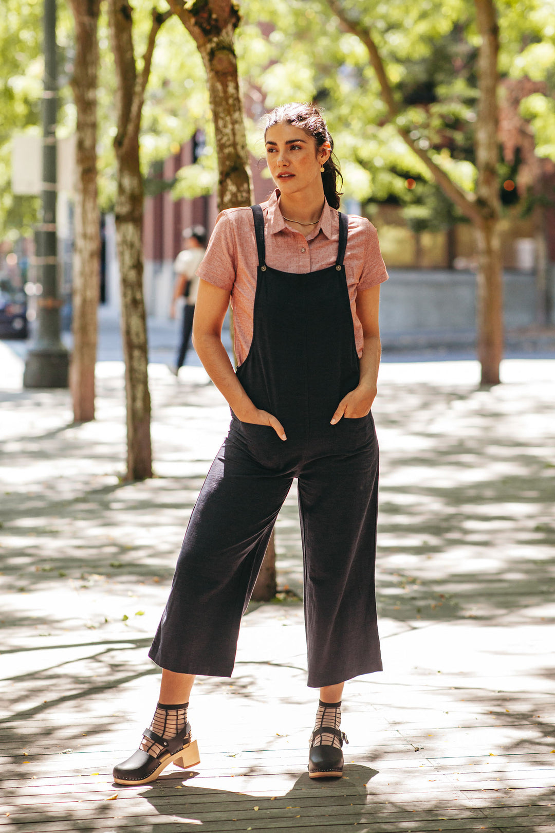 A woman stands outdoors on a sunny sidewalk, wearing Bridge & Burn’s Edie Overall in Navy with adjustable straps over a pink short-sleeve shirt and black heeled sandals, her hands in her pockets and green trees behind her.
