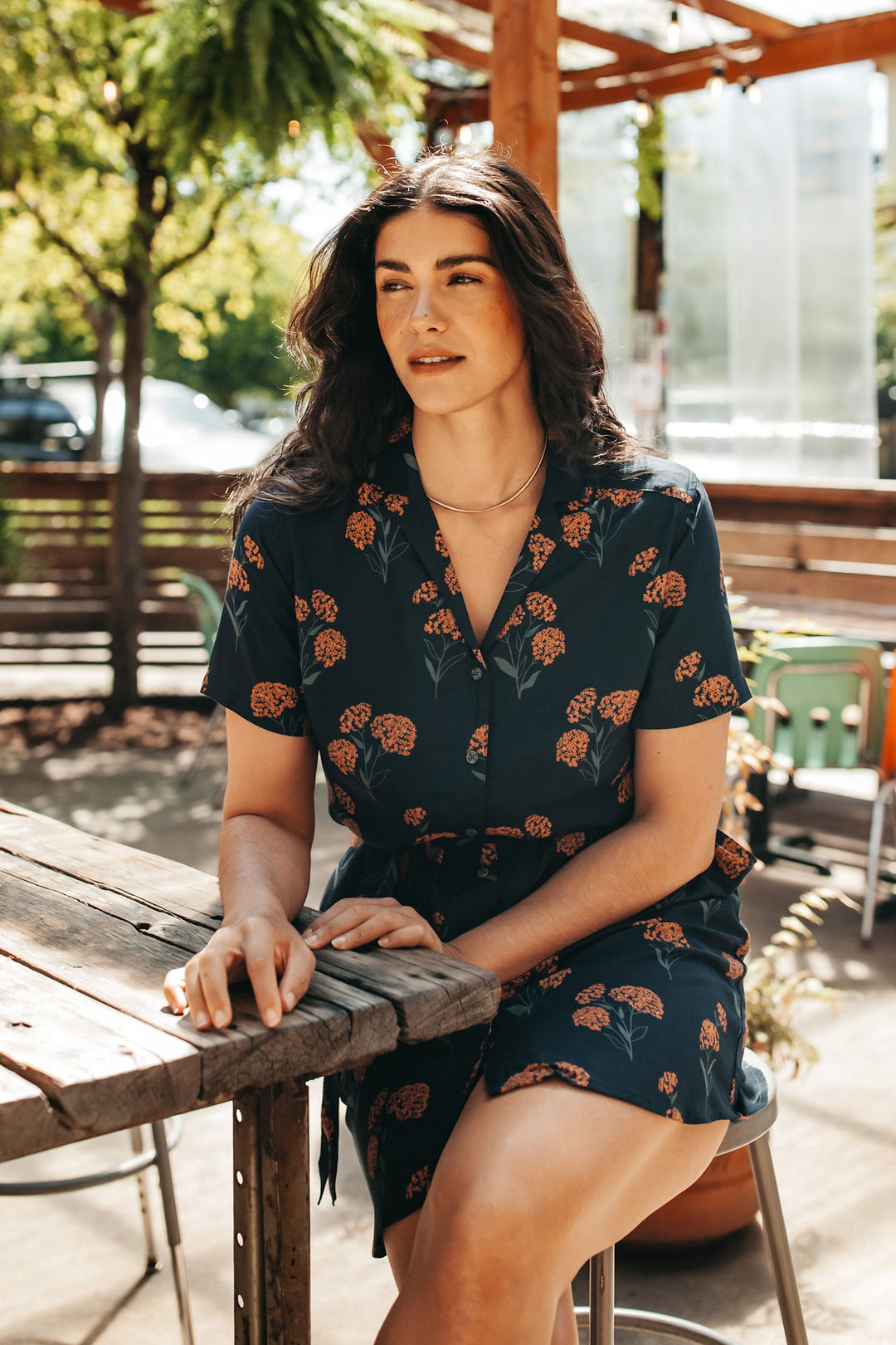 A woman with long dark hair relaxes on a wooden bench outdoors in a sunny, leafy setting, wearing the Bridge & Burn Sofia Shirt Dress in Wildflower. She sits with one arm on the table and has a gentle expression on her face.
