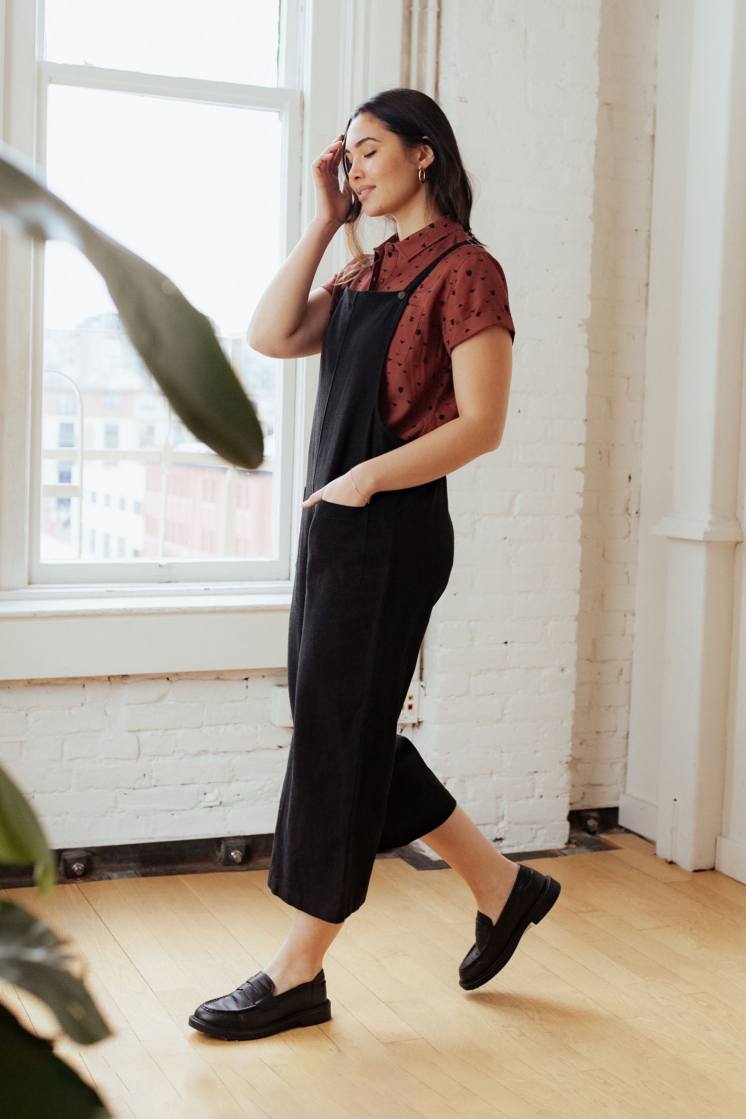 A woman stands indoors by a large window, smiling as she wears the Bridge & Burn Edie Overall in Black with a maroon polka dot blouse and black loafers. One hand touches her hair while the other rests in her pocket.