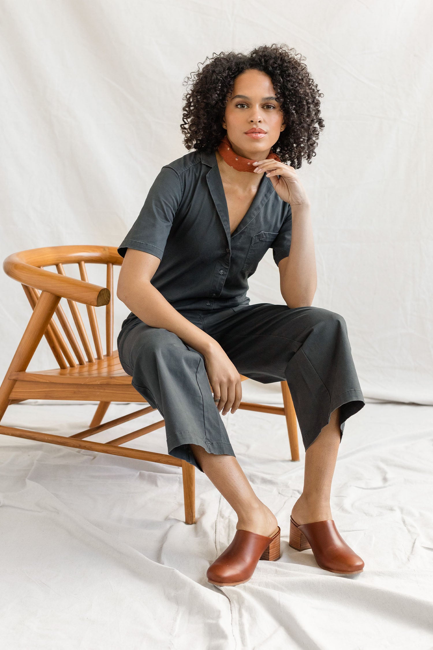 A person with curly hair sits on a wooden chair, wearing the Bridge & Burn Lou Utility Jumpsuit in Slate, paired with brown heeled clogs and a red bandana, set against a light, fabric-draped background.