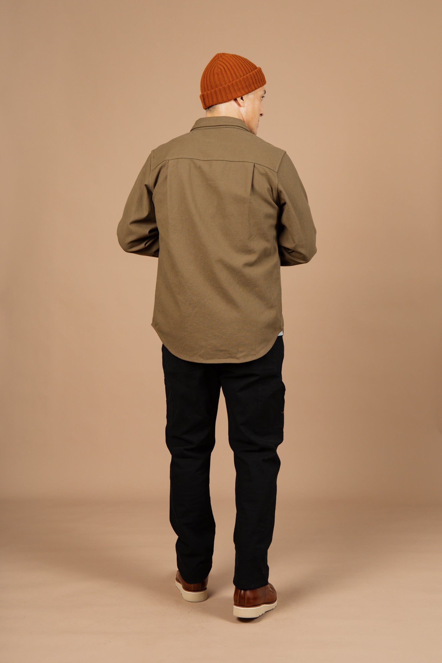 A person stands with their back to the camera, wearing the Bridge & Burn Bedford Shirt in Olive Twill, paired with black pants and brown shoes, set against a plain beige background.