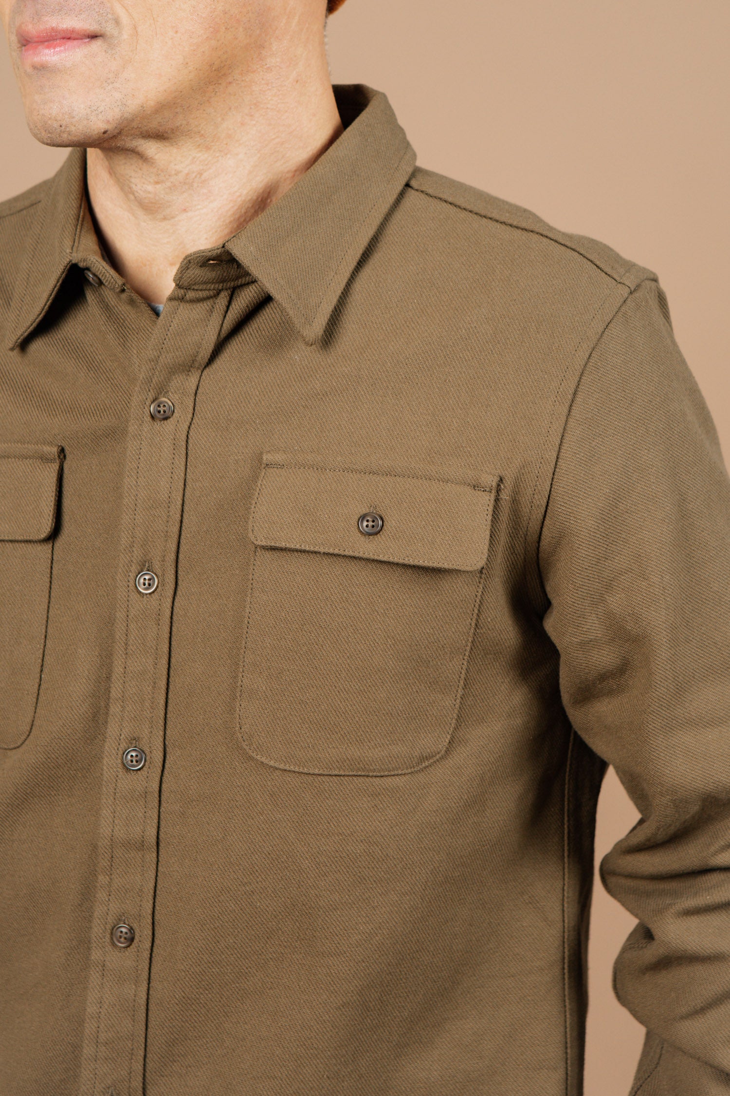 A man wearing the Bridge & Burn Bedford Shirt in Olive Twill, featuring two front pockets, stands against a beige background with only his lower face and upper torso visible.