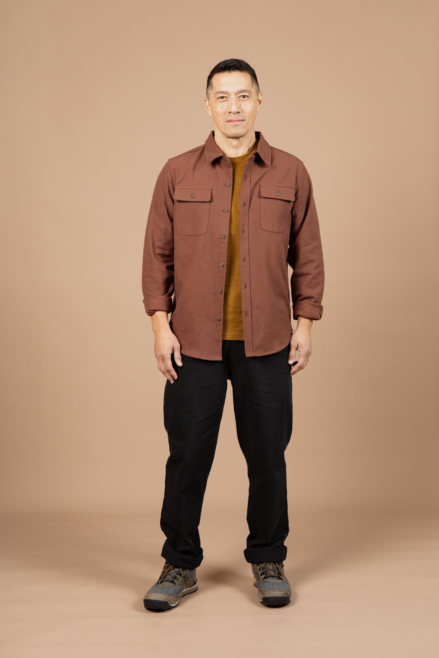 A man faces forward against a beige background, wearing Bridge & Burn’s Bedford Shirt in Penny Twill over a mustard shirt, paired with black pants and gray hiking shoes.