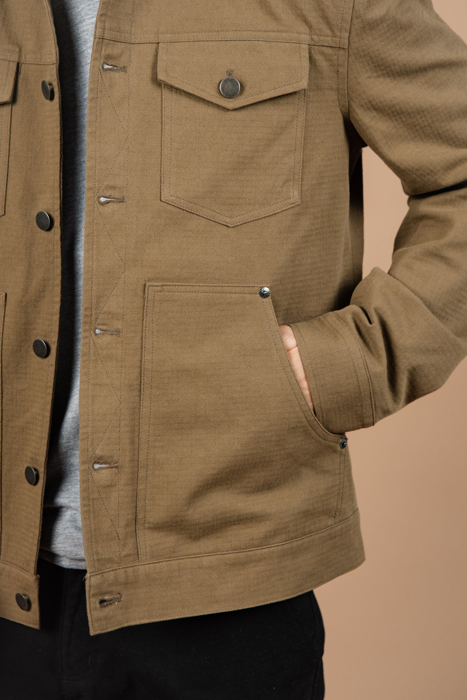 A person wears the Bridge & Burn Burl Trucker Jacket in Warm Olive, featuring a flap chest pocket and side hand pocket, standing against a beige background with their right hand in the side pocket. Only the torso and part of the arms are shown.