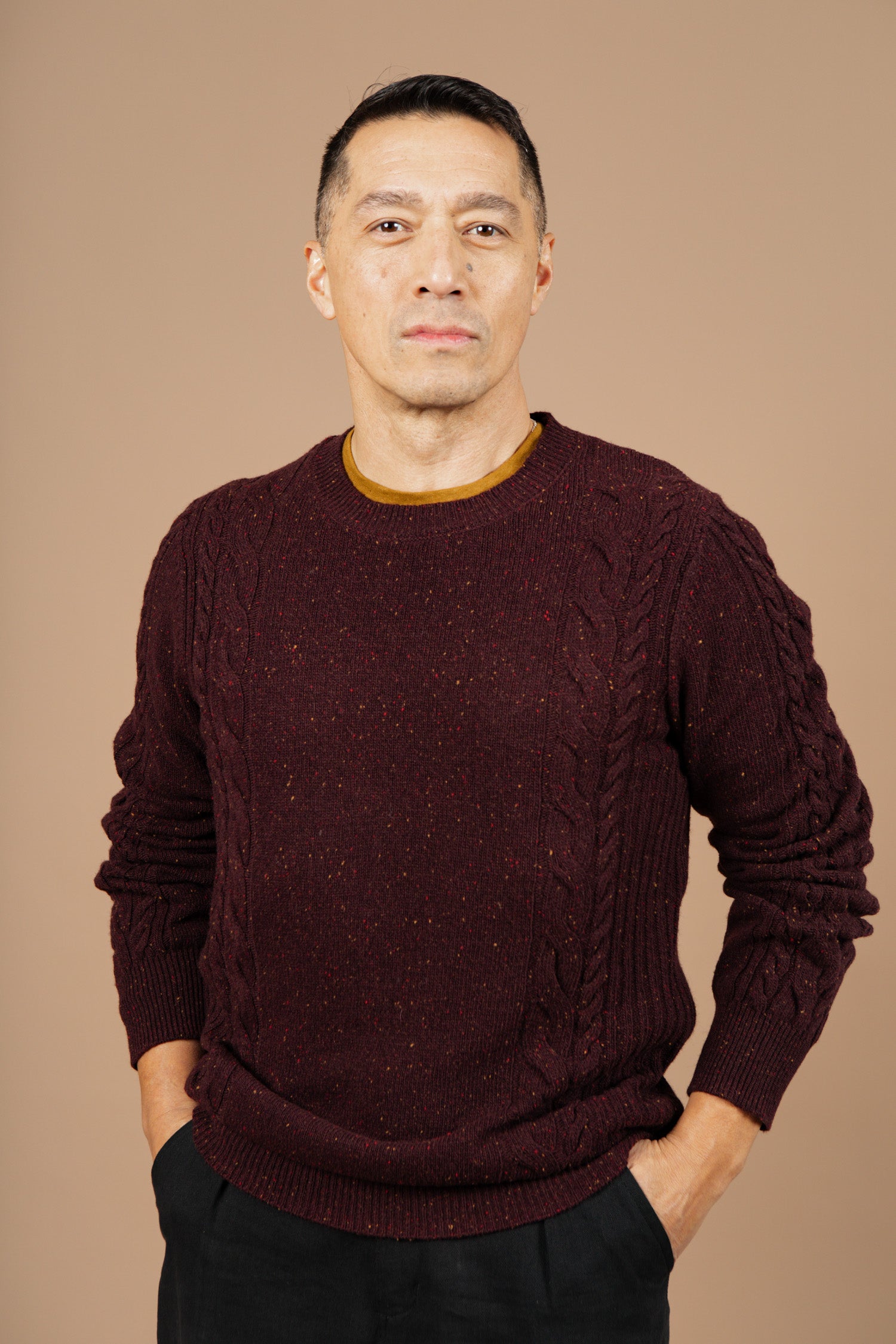 A man with short dark hair stands facing the camera against a plain beige background, wearing the Bridge & Burn Hugo Sweater in Burgundy Donegal with dark pants and his hands in his pockets.
