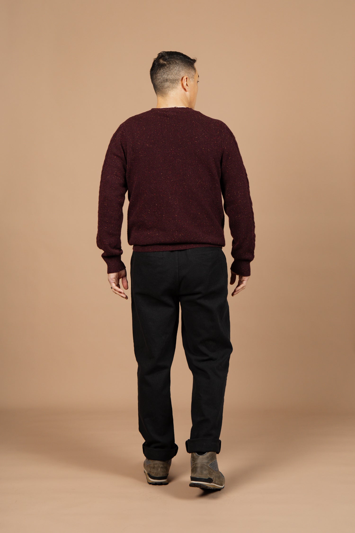 Facing away from the camera against a beige background, a person with short hair wears the Bridge & Burn Hugo Sweater in Burgundy Donegal, paired with black pants and brown shoes.
