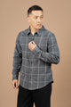 A man wearing the Bridge & Burn Larson Shirt in Shadow Twill Windowpane and black pants stands against a plain beige background, looking to his right while adjusting his shirt with one hand.