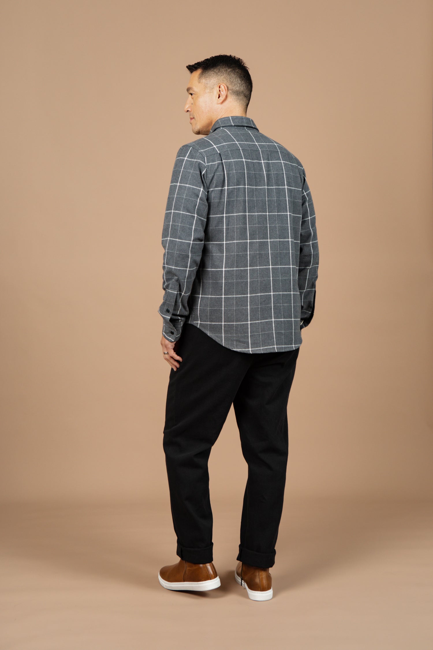 A man stands facing away, wearing the Bridge & Burn Larson Shirt in Shadow Twill Windowpane, black pants, and brown sneakers against a plain beige background.