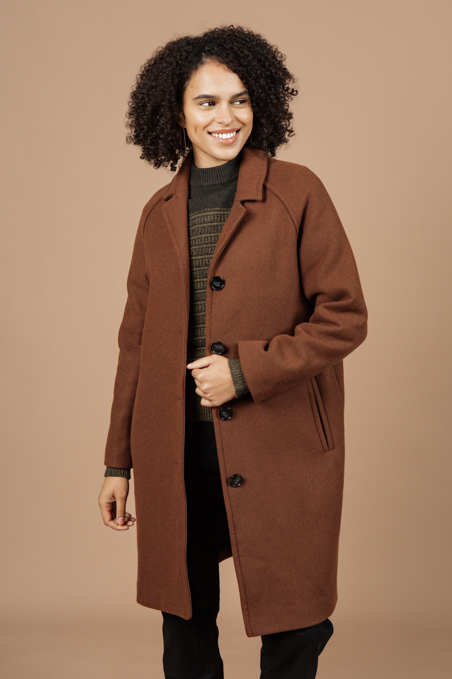 A person with curly hair smiles while wearing the Bridge & Burn Alberta Car Coat in Fired Brick over a green sweater and black pants, standing against a plain light brown background.