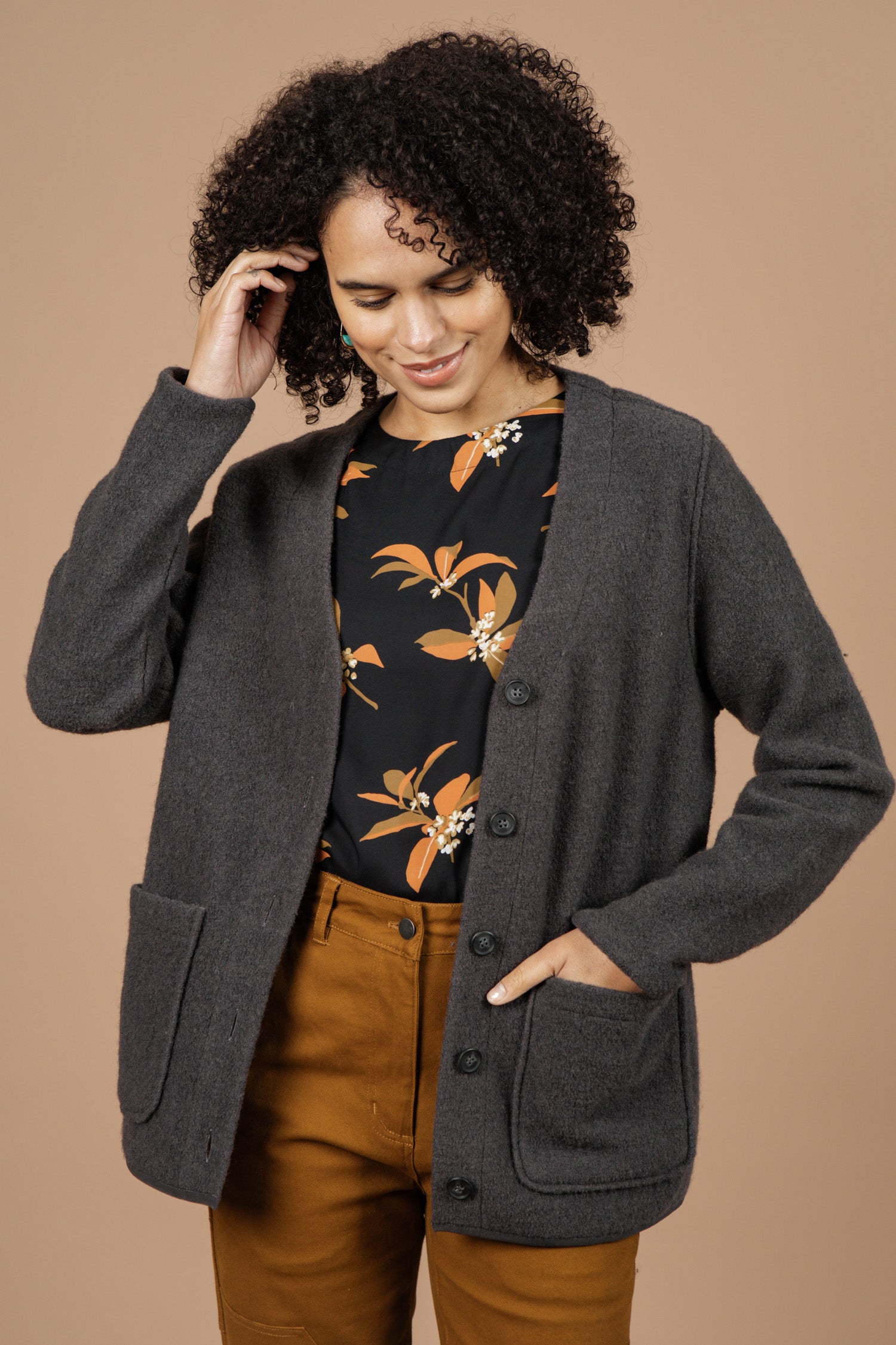 A woman with curly hair, wearing the Bridge & Burn Whit Cardigan in Charcoal over a black shirt with an orange floral pattern and brown pants, stands smiling against a beige background and touches her hair.