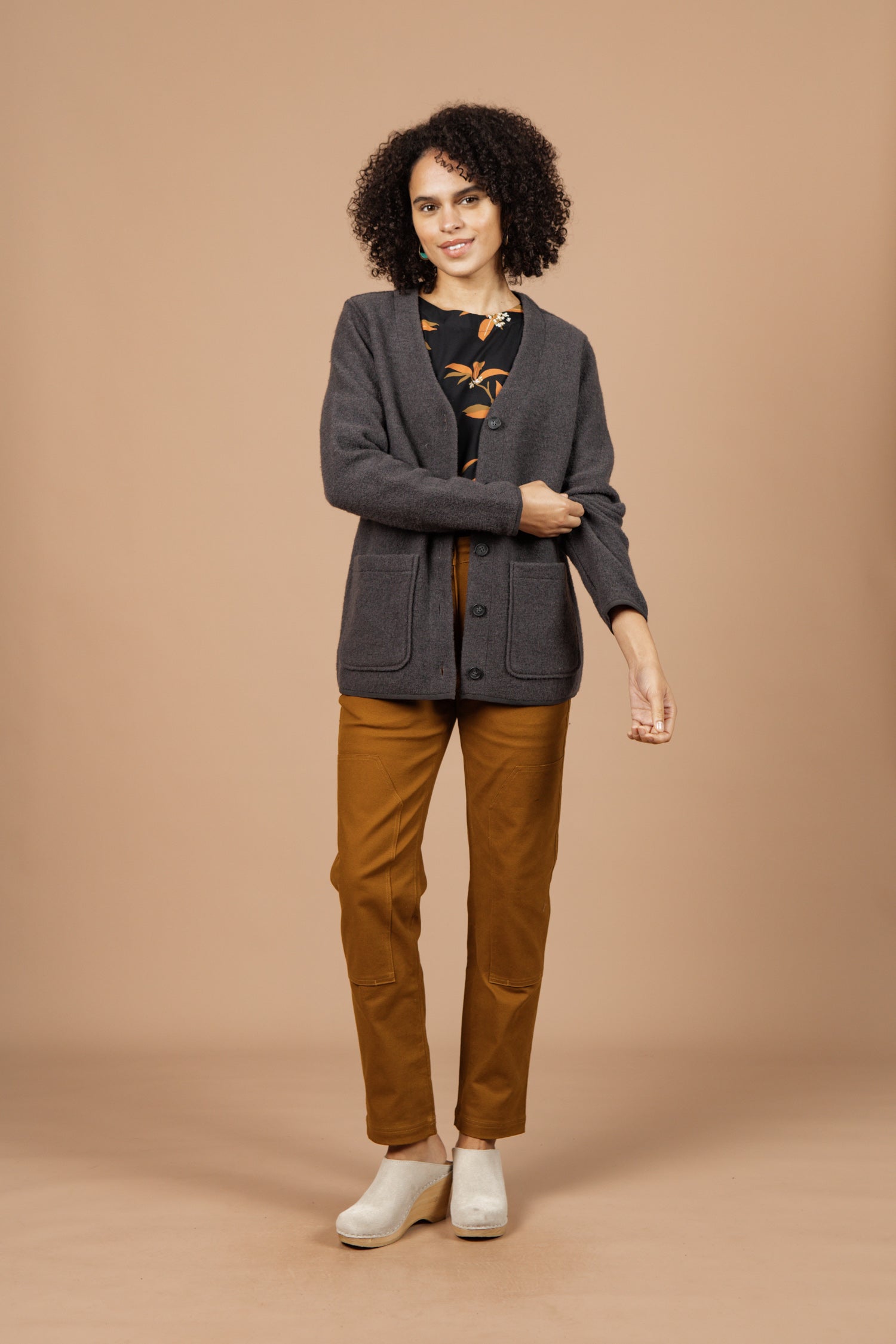 A woman with curly hair smiles against a beige background, wearing the Bridge & Burn Whit Cardigan in Charcoal over a patterned black top, mustard pants, and light shoes. She holds one arm with her other hand.
