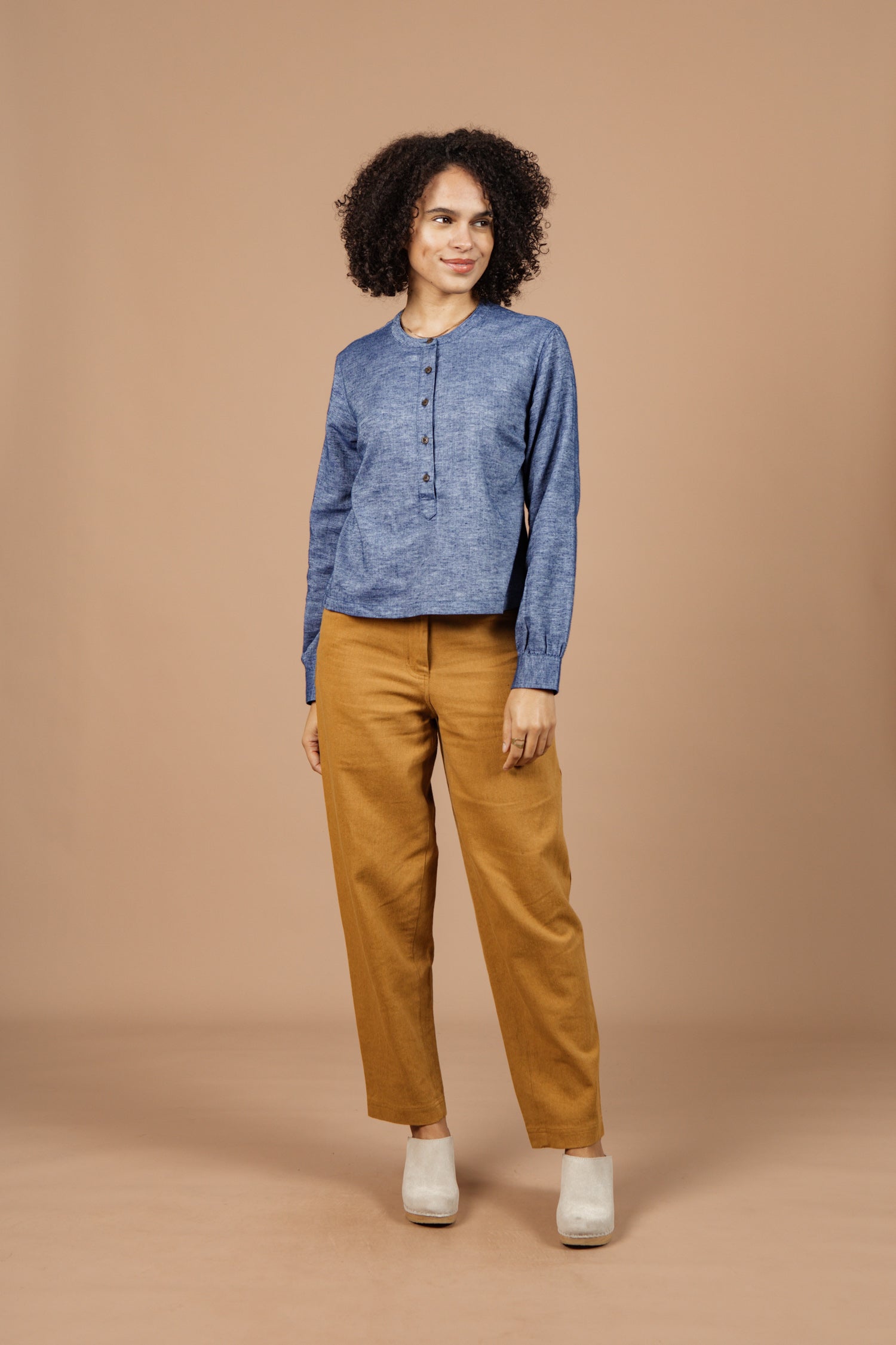 A person with curly hair stands against a beige background, smiling slightly and looking to the side, dressed in the Bridge & Burn Yarra Pullover in Indigo Herringbone—showcasing stylish, sustainable fashion.