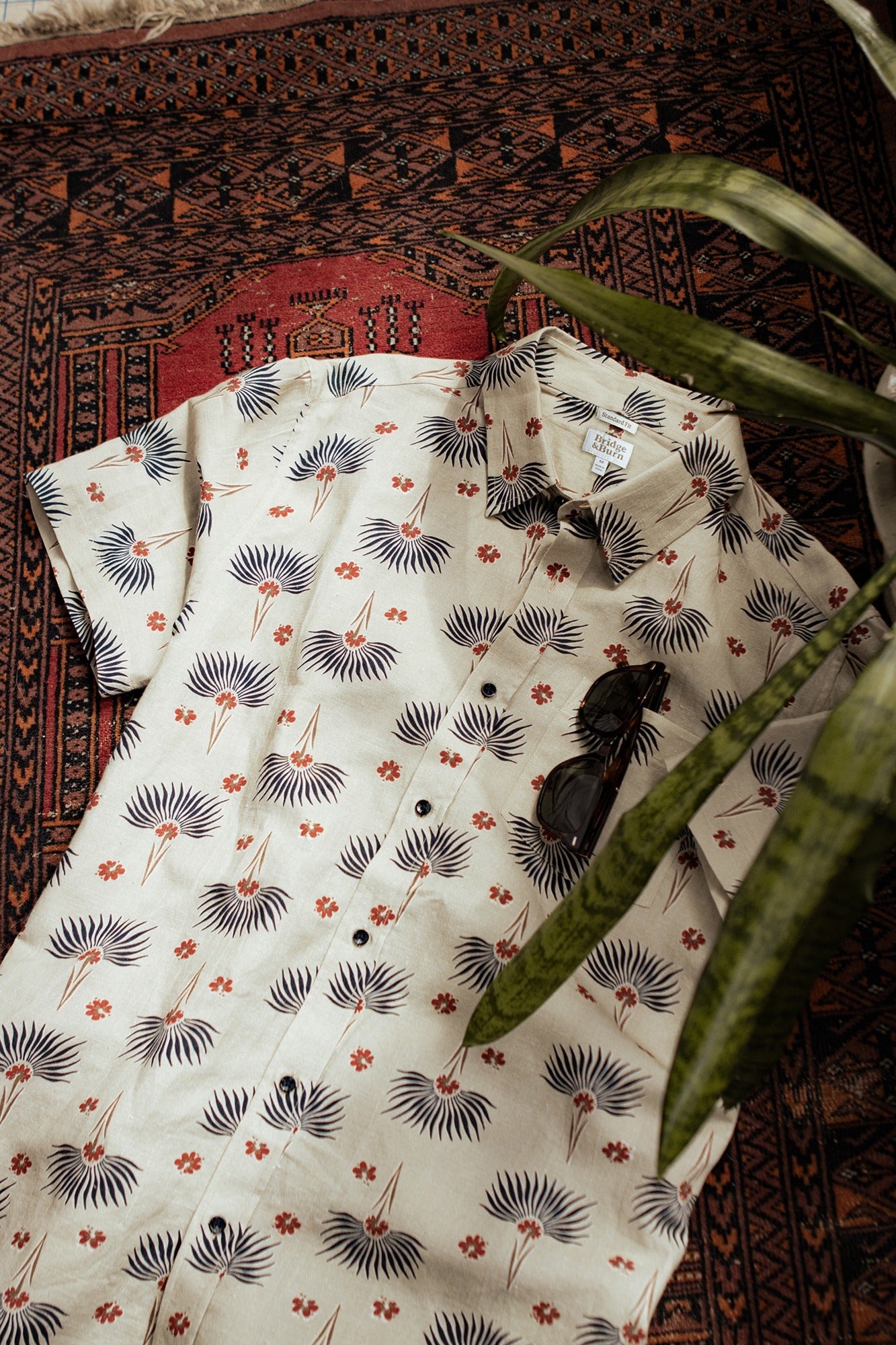 The Bridge & Burn Bea Shirt / Aloha Floral, a classic short-sleeve button down with white, blue, and orange floral print, is styled on a patterned rug with dark sunglasses and a green plant leaf in the foreground.
