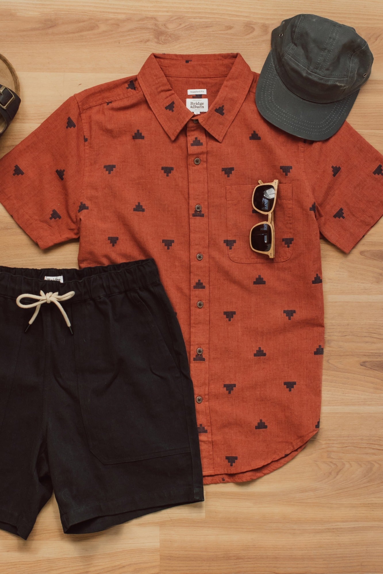 A flat lay of Bridge & Burn's Malcolm Shirt in Rust Fil-Coupé Pyramids, paired with black drawstring shorts, a dark green cap, and sunglasses, all arranged on a wooden floor.