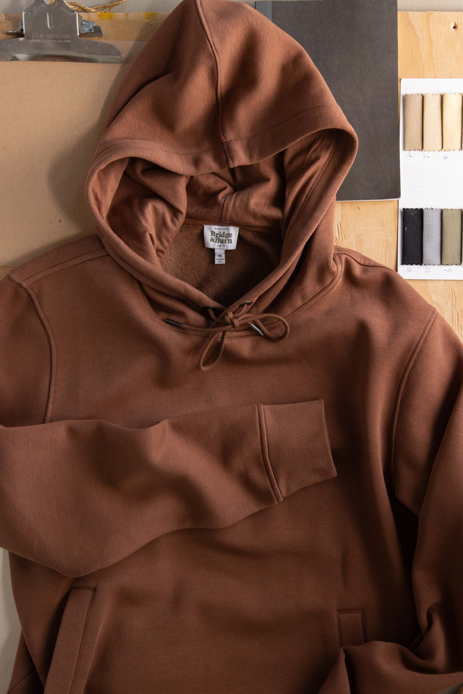The Williams Pullover Hoodie in Burnt Umber by Bridge & Burn rests on a wooden surface beside fabric swatches, a clipboard, and paper. This organic cotton sweatshirt features a front pocket and hood with drawstring for eco-friendly comfort.