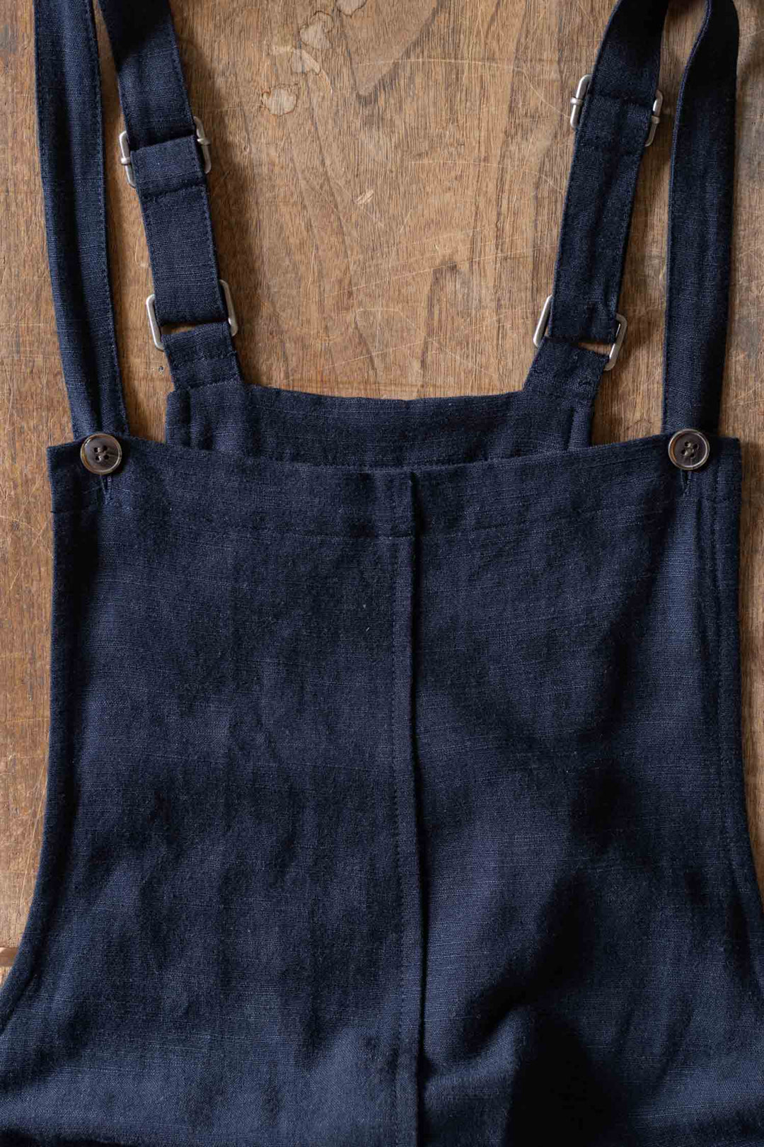 The Edie Overall in Navy from Bridge & Burn features a wide-leg silhouette with adjustable shoulder straps and button details, crafted from a soft linen-cotton blend and displayed flat on a wooden surface.