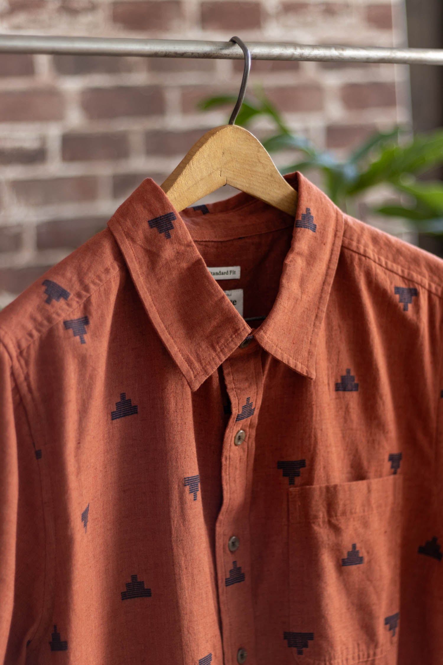 The Malcolm Shirt in Rust Fil-Coupé Pyramids by Bridge & Burn, a sustainable button-up with a dark geometric pattern, hangs on a wooden hanger against a brick wall background.