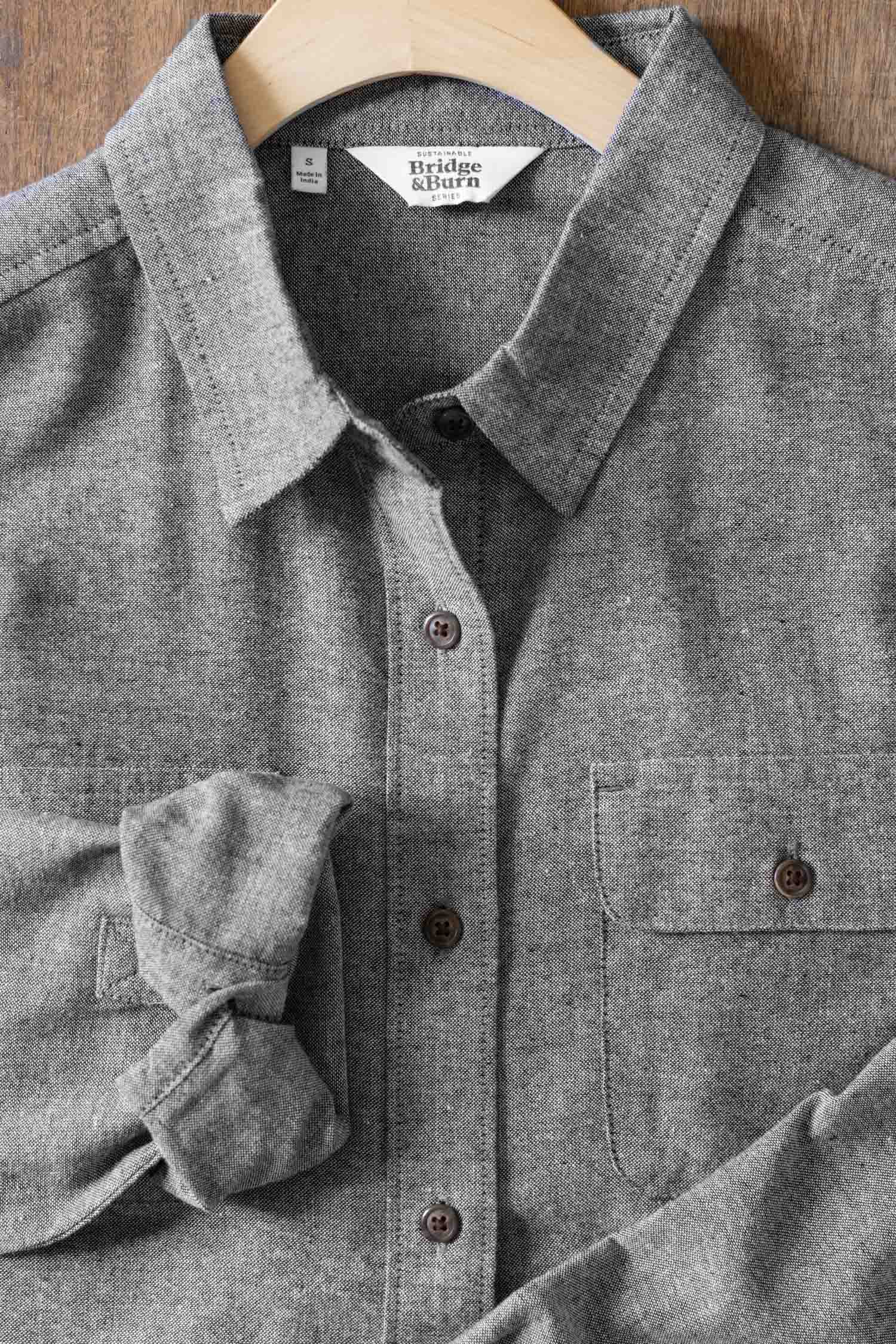 The Lenon Shirt in Iron Chambray by Bridge & Burn is shown on a wooden hanger, with one sleeve partially rolled up and set on a wooden surface. This relaxed fit gray shirt features a front pocket.