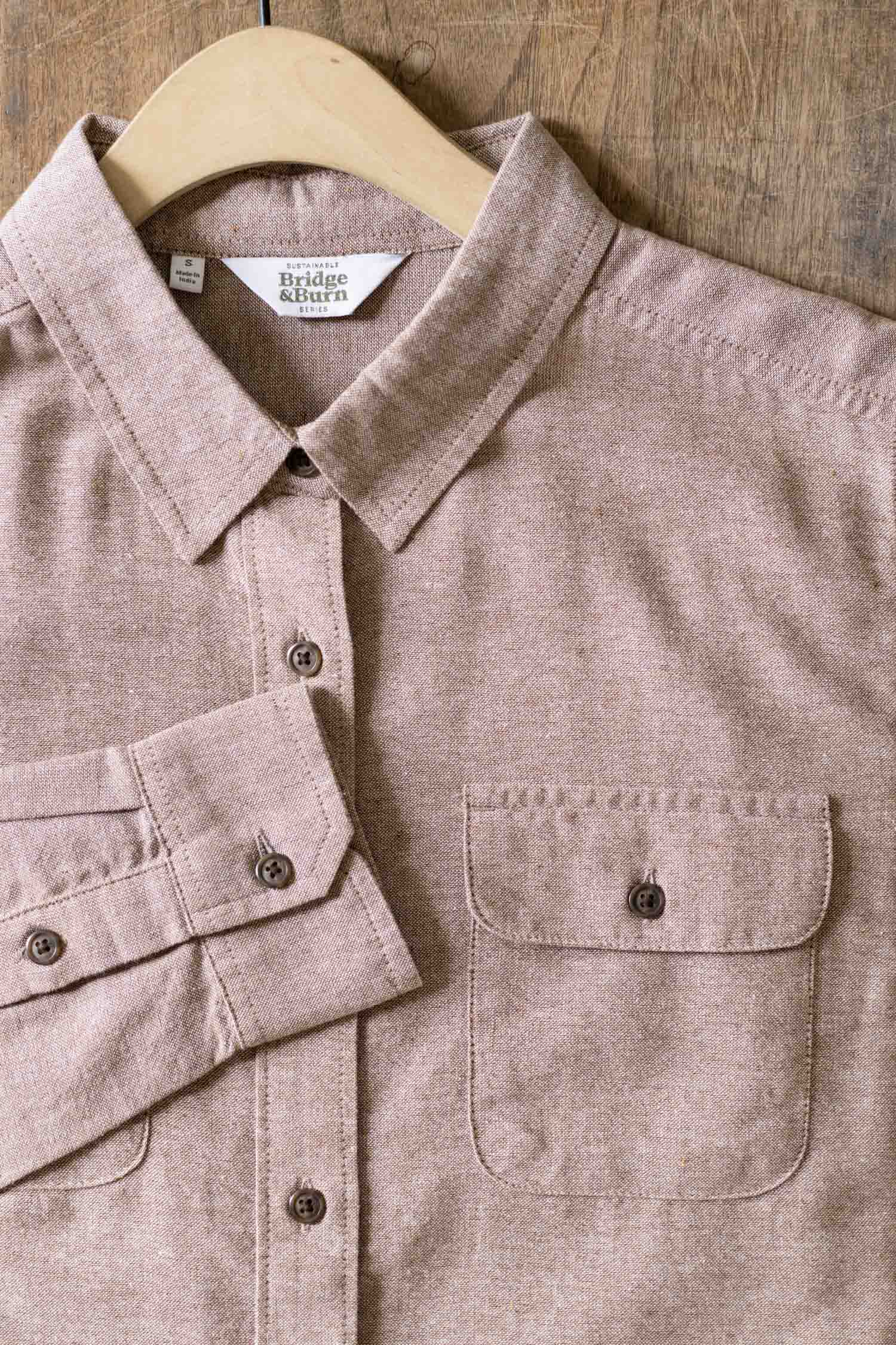 The Bridge & Burn Lenon Shirt in Rust Chambray is a light brown, long-sleeve relaxed fit shirt with a single chest pocket, crafted from recycled cotton with a subtly textured look, shown on a wooden hanger against a wood background.