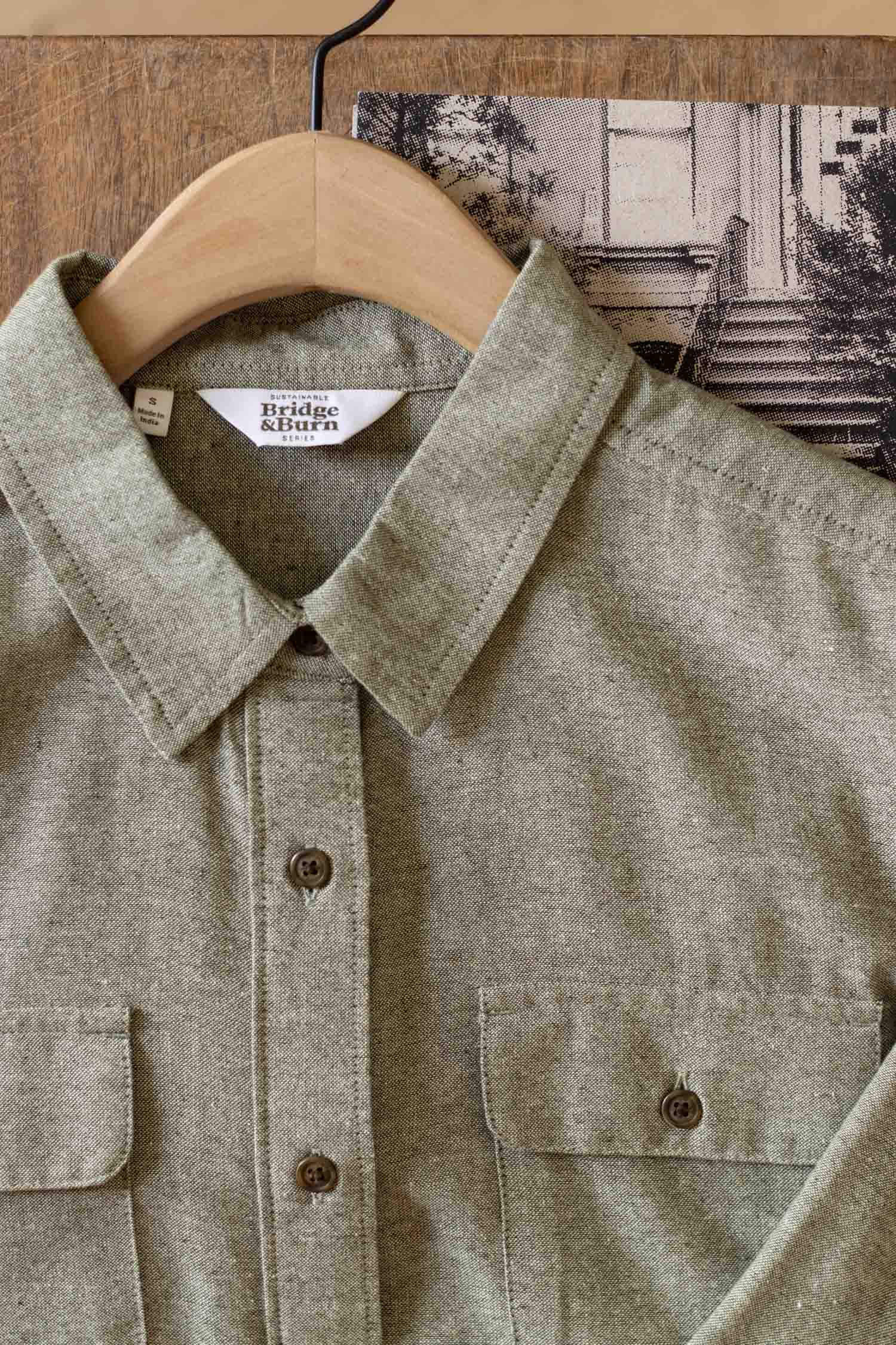 A close-up of the Bridge & Burn Lenon Shirt in Sage Chambray—made from recycled cotton—displays its collar, chest pockets, and buttons on a wooden hanger, with a black-and-white photo partially visible in the background.
