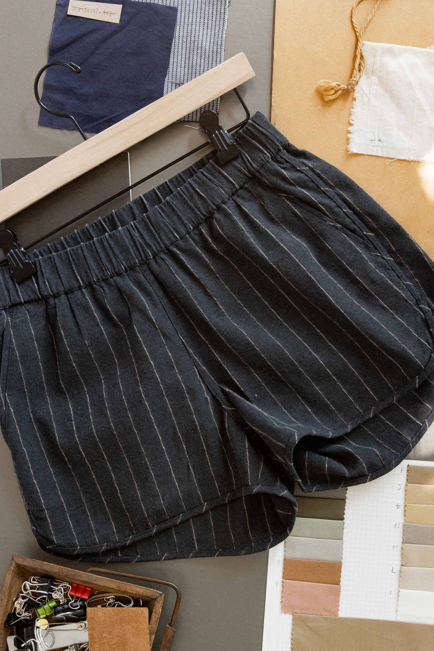 The Bridge & Burn Luca Track Short in Charcoal Stripe hangs on a black hanger, surrounded by fabric swatches, paper, and small office supplies on a tabletop.