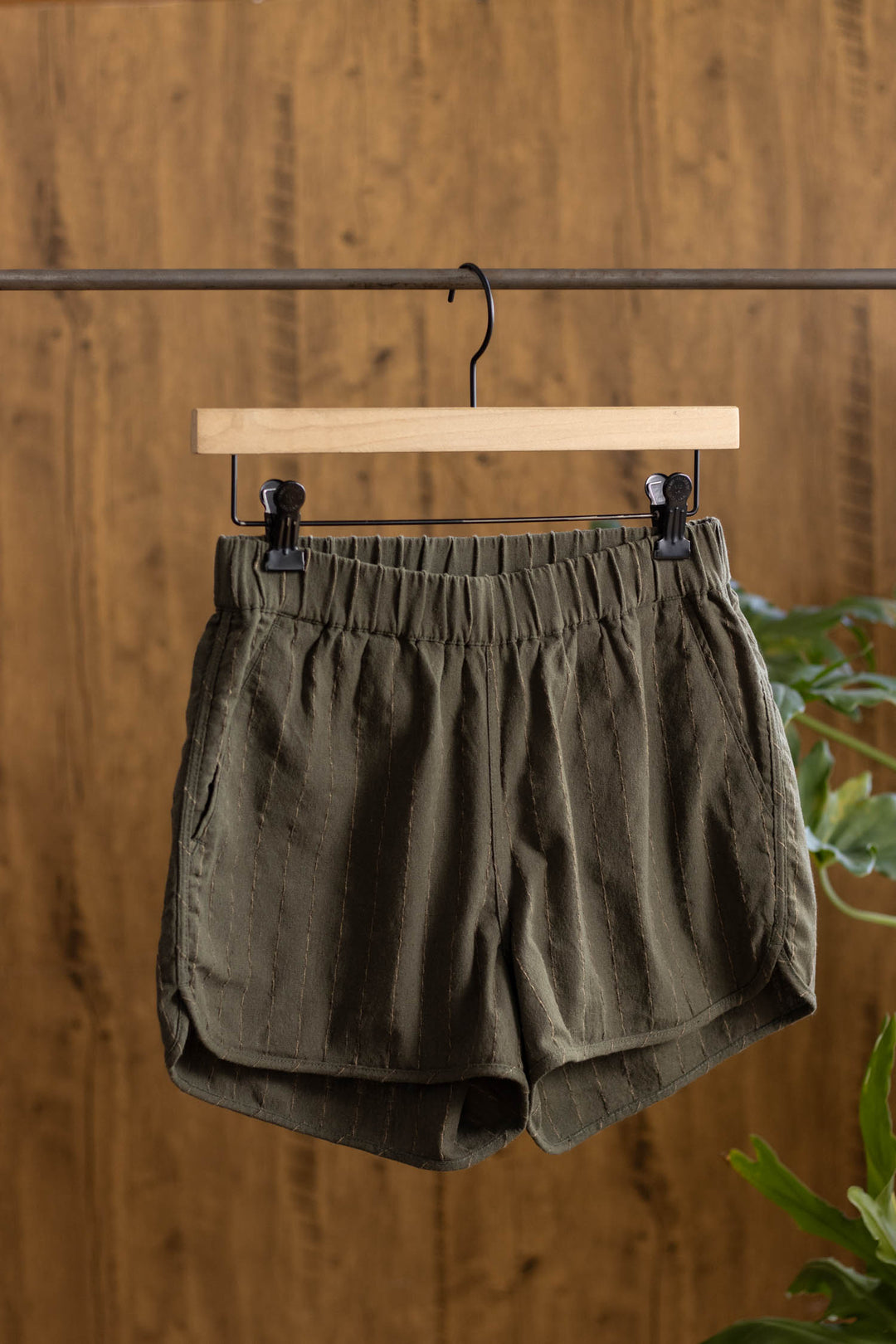 The Bridge & Burn Luca Track Short in Olive Stripe, made from cotton-linen with an elastic waistband, hangs on a wooden hanger against a wooden background with green plants partially visible on the right.