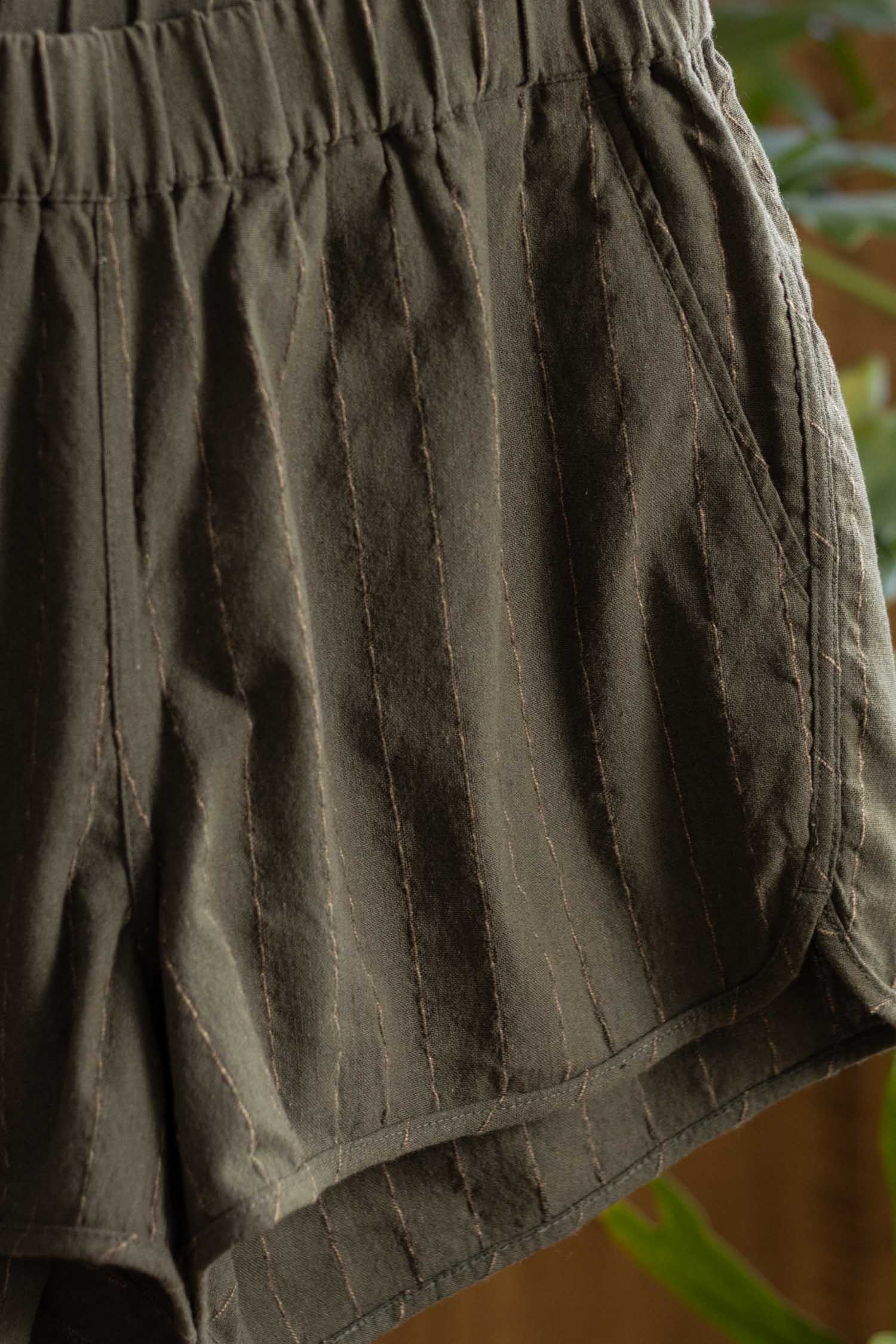 Close-up of the Luca Track Short in Olive Stripe by Bridge & Burn, featuring soft cotton-linen fabric with subtle vertical pinstripes, an elastic waistband, and side pocket. The textured material stands out against a blurred green background.
