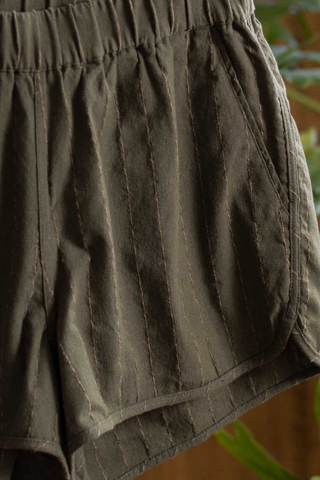 Close-up of the Luca Track Short in Olive Stripe by Bridge & Burn, featuring soft cotton-linen fabric with subtle vertical pinstripes, an elastic waistband, and side pocket. The textured material stands out against a blurred green background.