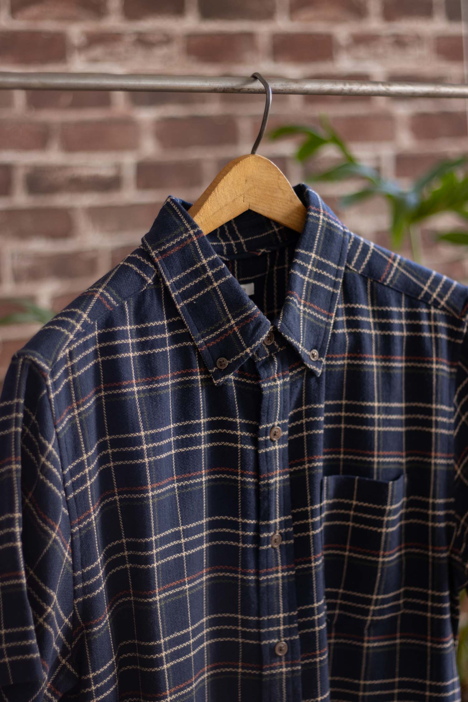 The Bridge & Burn Jordan Slim Shirt in Dusk Herringbone Plaid, featuring modern navy blue with white, beige, and red lines, hangs on a wooden hanger against a blurred brick wall background.