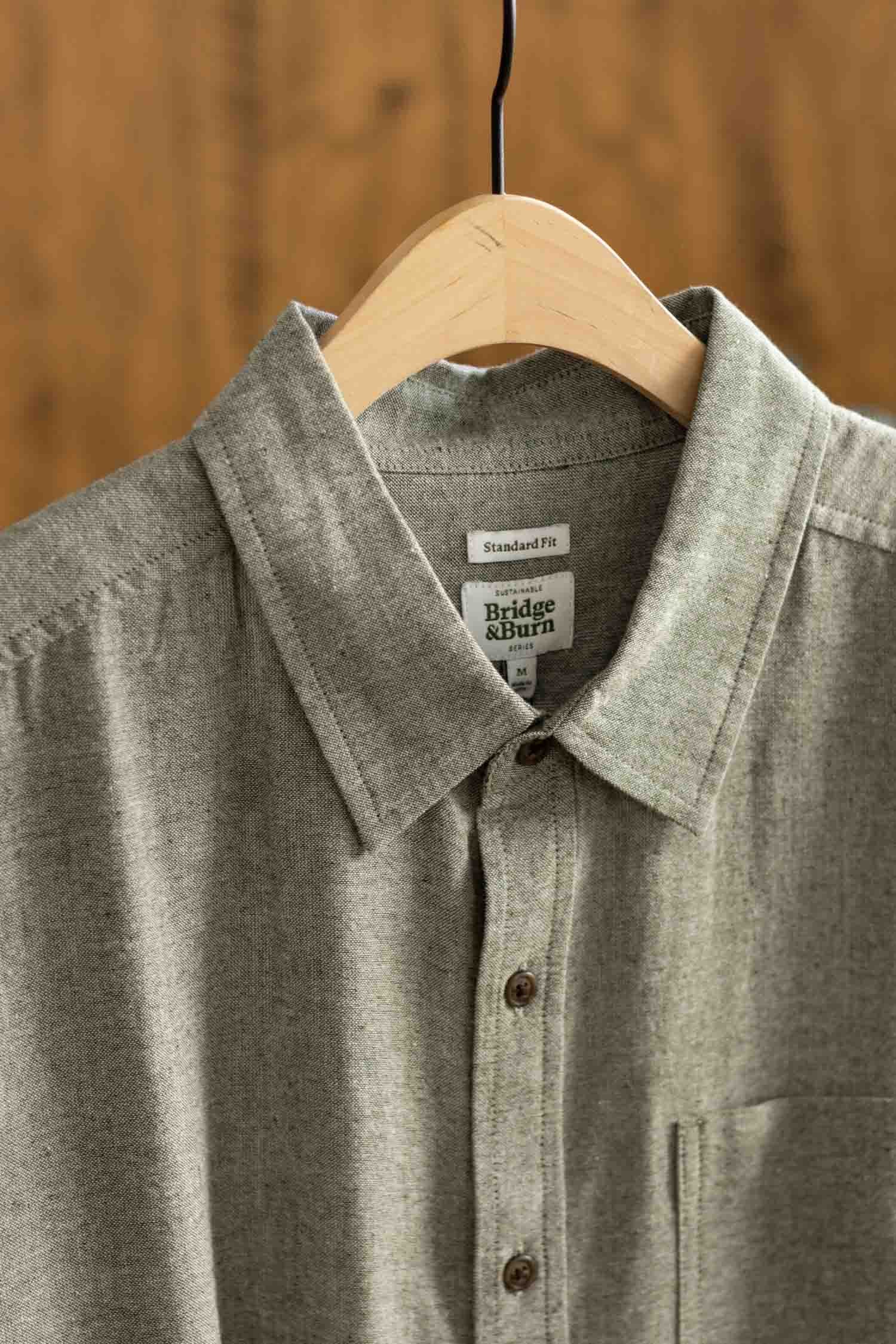 A sage chambray Larson Shirt by Bridge & Burn hangs on a wooden hanger. Featuring a classic fit and button-up style, this men's shirt is made from recycled cotton and stands out against a blurred warm brown background.
